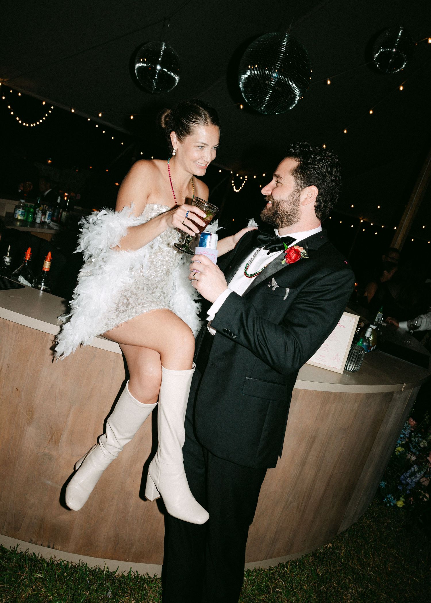 Bride in feathered dress sitting on bar counter toasting with groom in tuxedo.