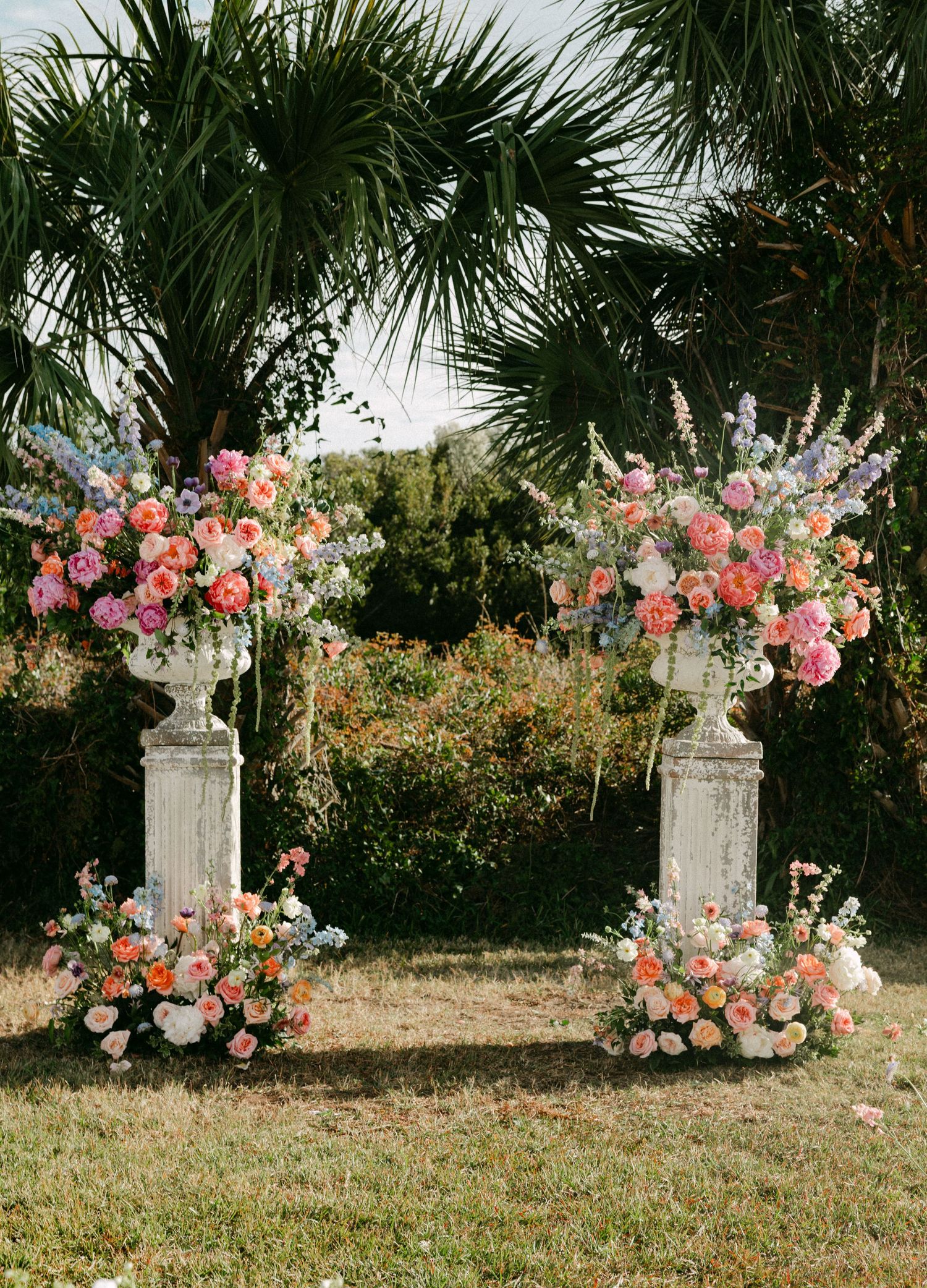 Elegant outdoor wedding altar with white stone urns overflowing with pink, coral, and blue flowers.