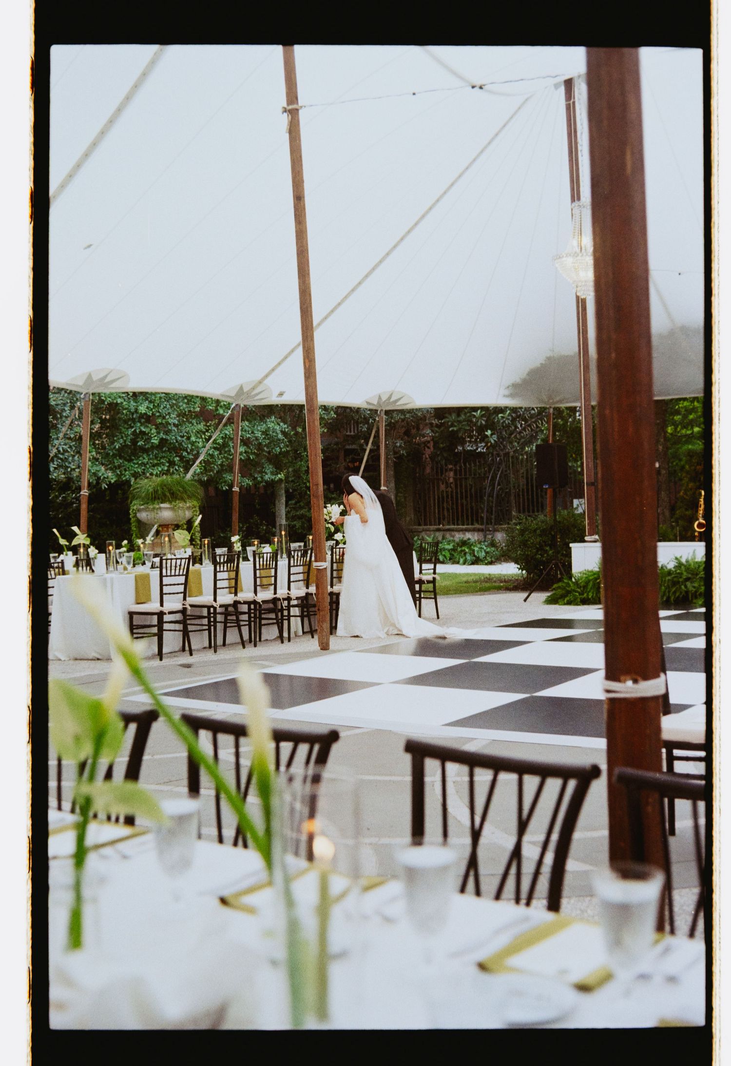 Bride in white gown and veil standing on black and white checkered dance floor under tent.