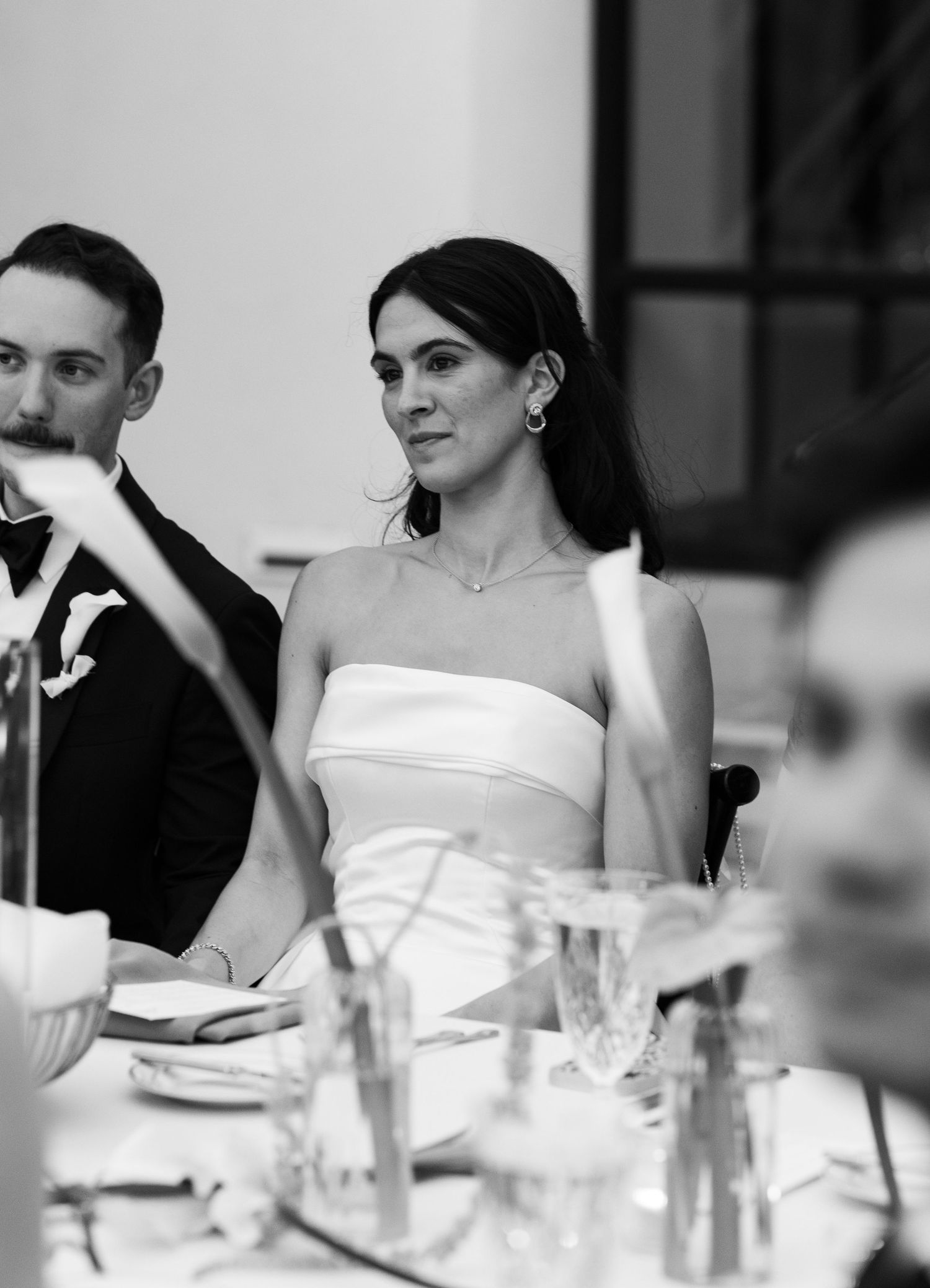 Black and white photo of a bride in a strapless white gown seated at a wedding reception table.