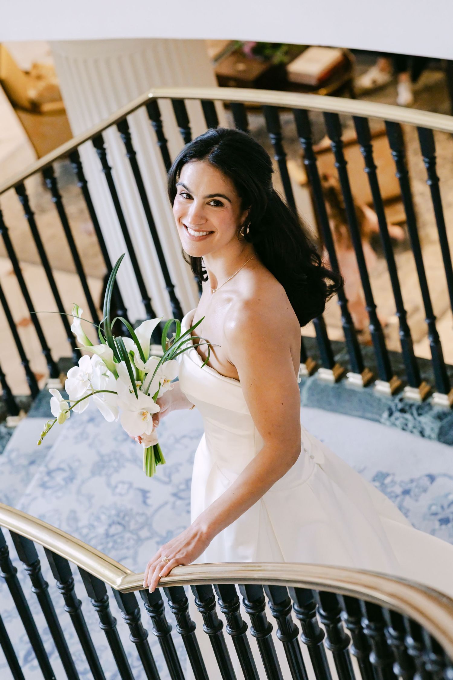 Smiling dark-haired bride in strapless white gown holding white calla lily bouquet on elegant spiral staircase.