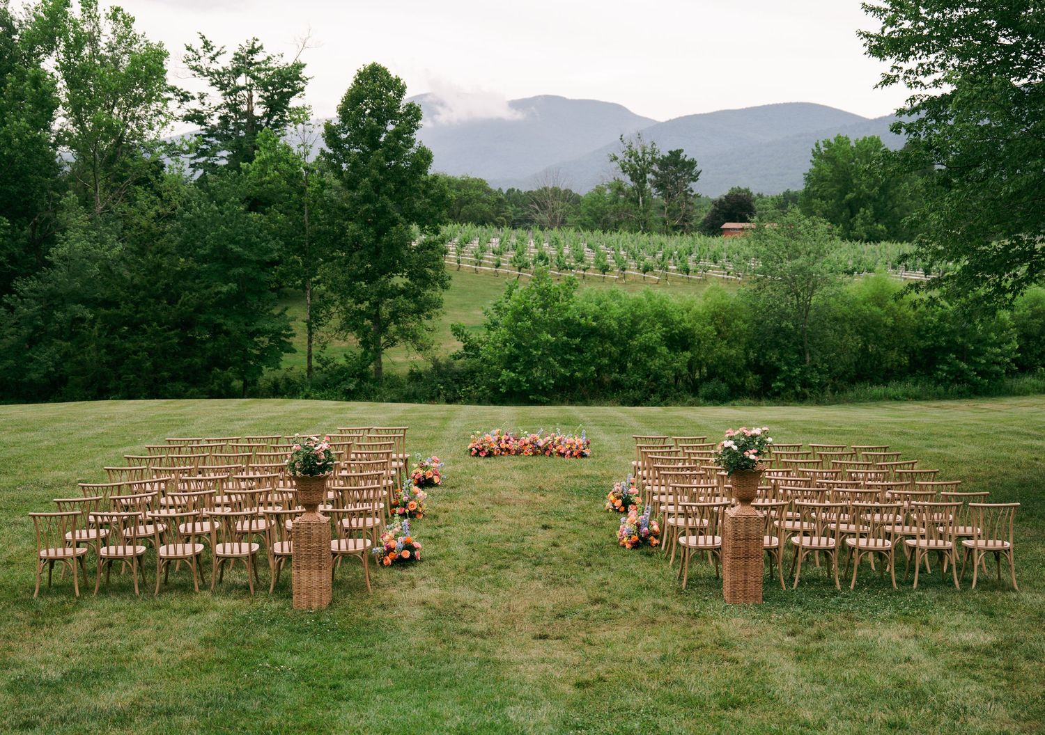 Outdoor wedding ceremony setup with wooden chairs on green lawn, floral arrangements, and mountain vineyard backdrop.
