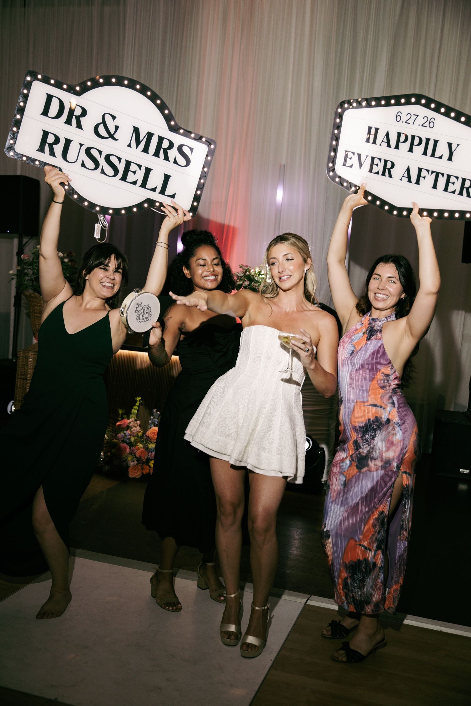 Bride in white mini dress poses with three friends holding DR & MRS RUSSELL marquee signs at reception.