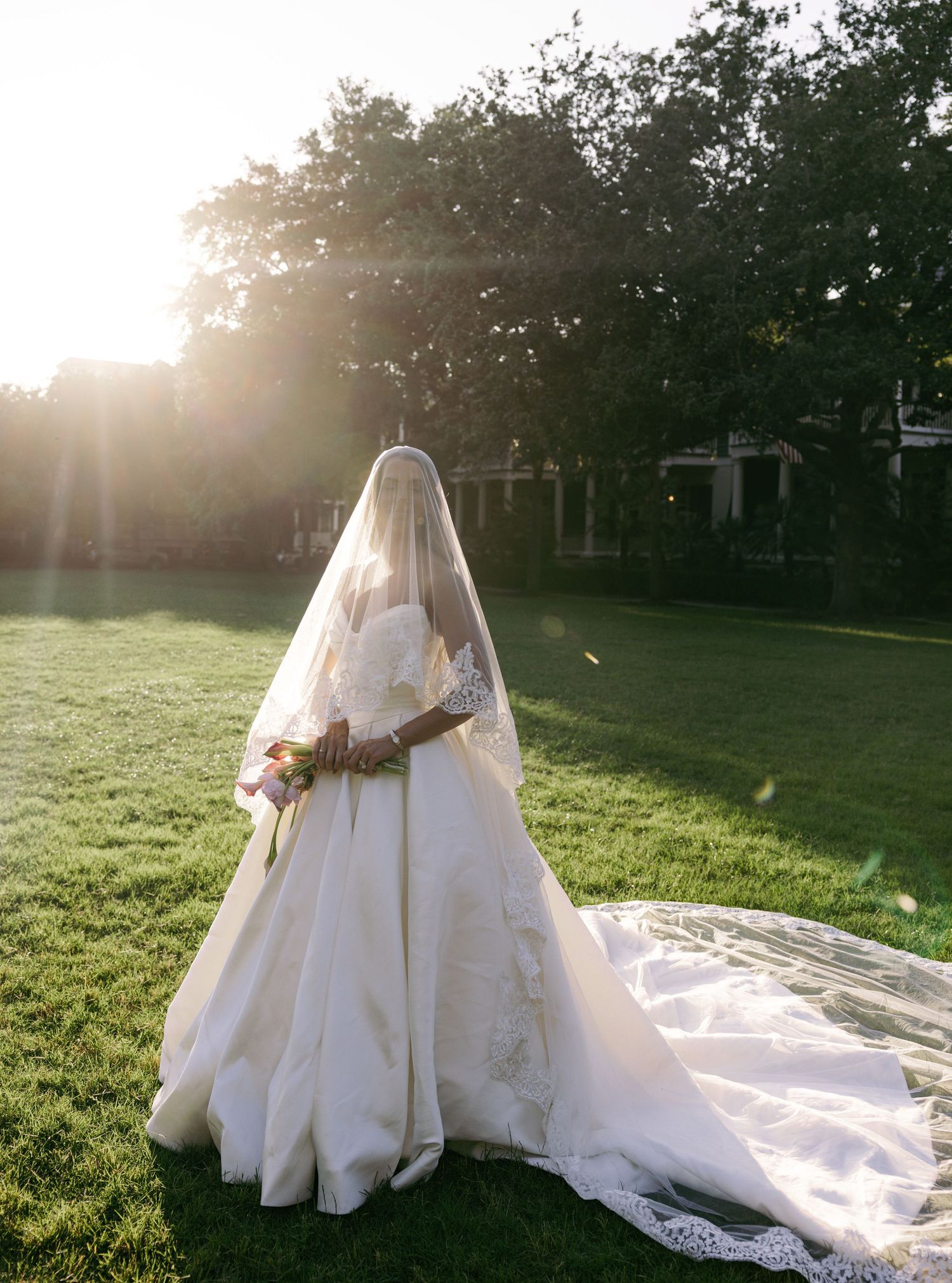 Bride in elegant ball gown with lace veil and train stands on green lawn at golden hour sunset.