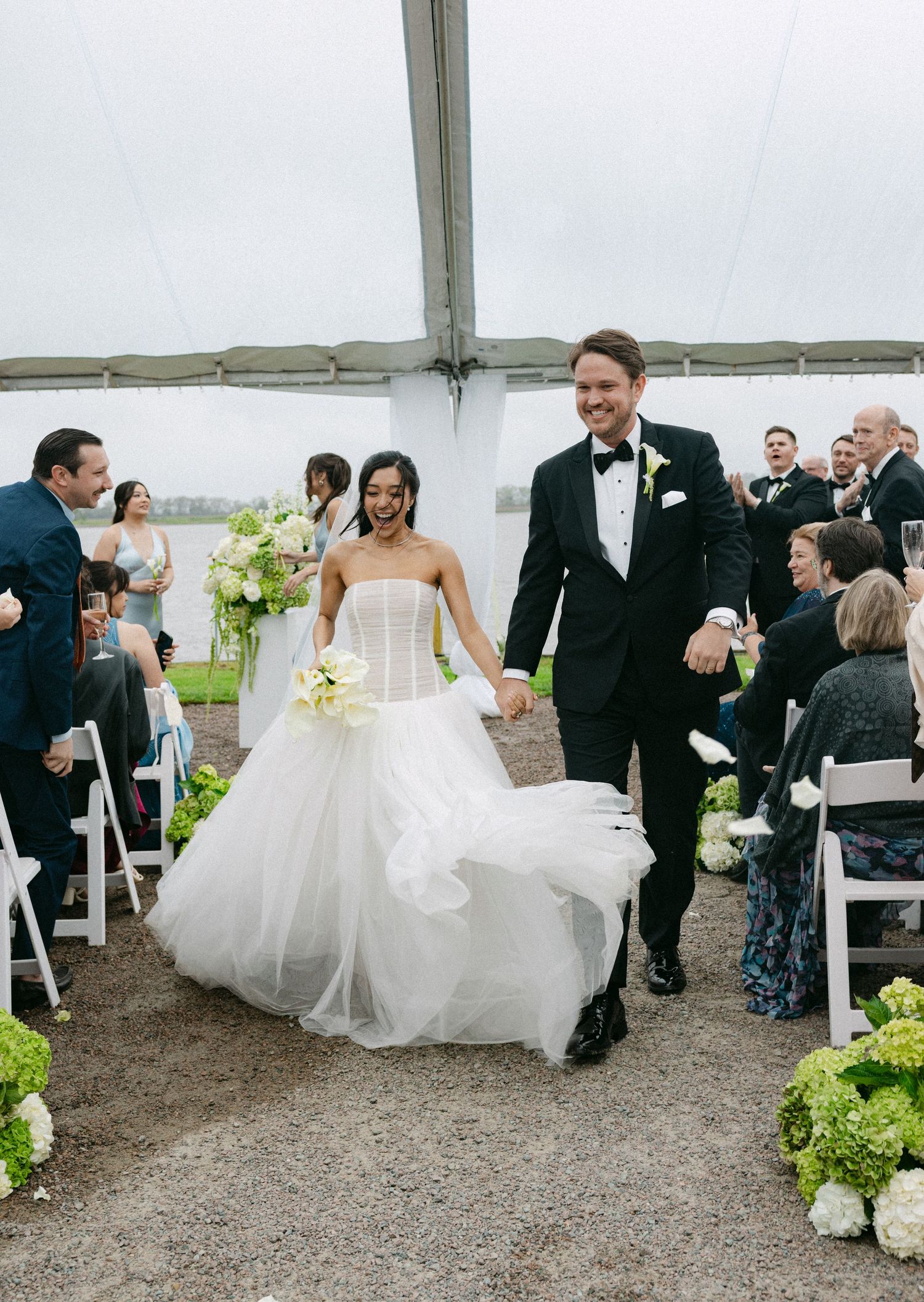 Bride and groom walk down the aisle hand in hand under a tent, surrounded by guests and green floral arrangements.