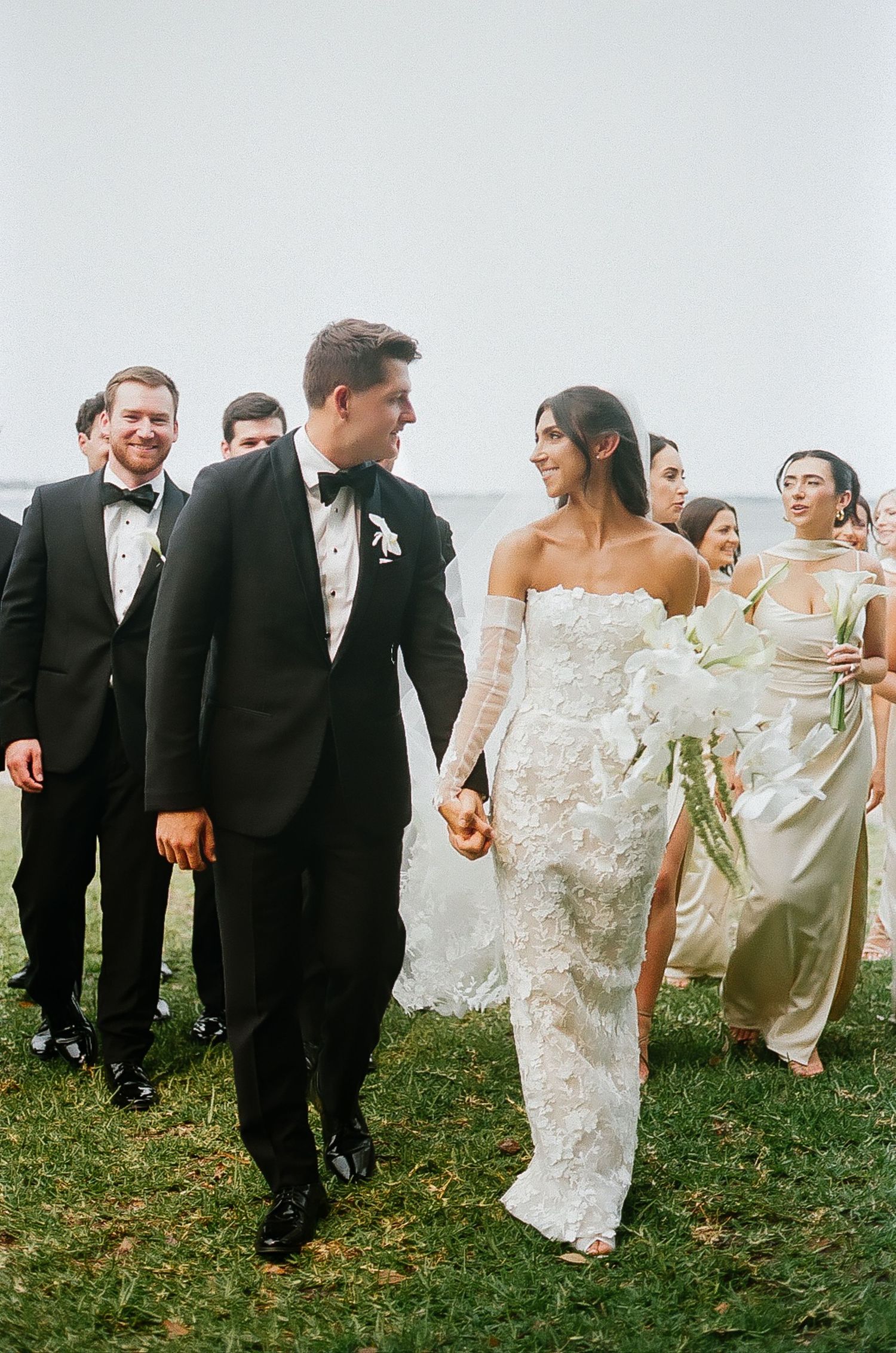 Wedding party in formal attire walks and laughs together on grassy lawn.