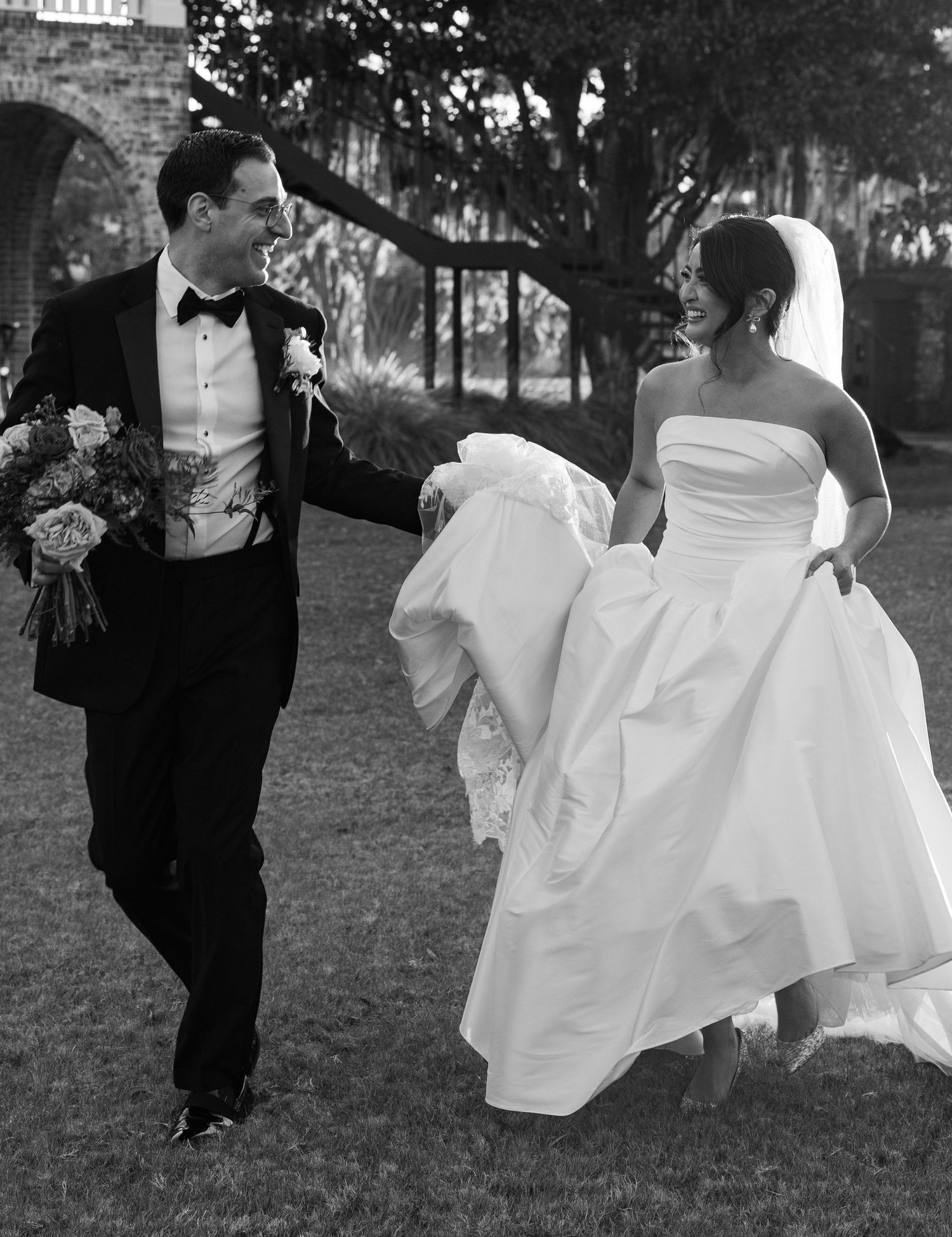 A bride in a flowing white gown and groom in classic tuxedo share a joyful moment while walking on grass outdoors.