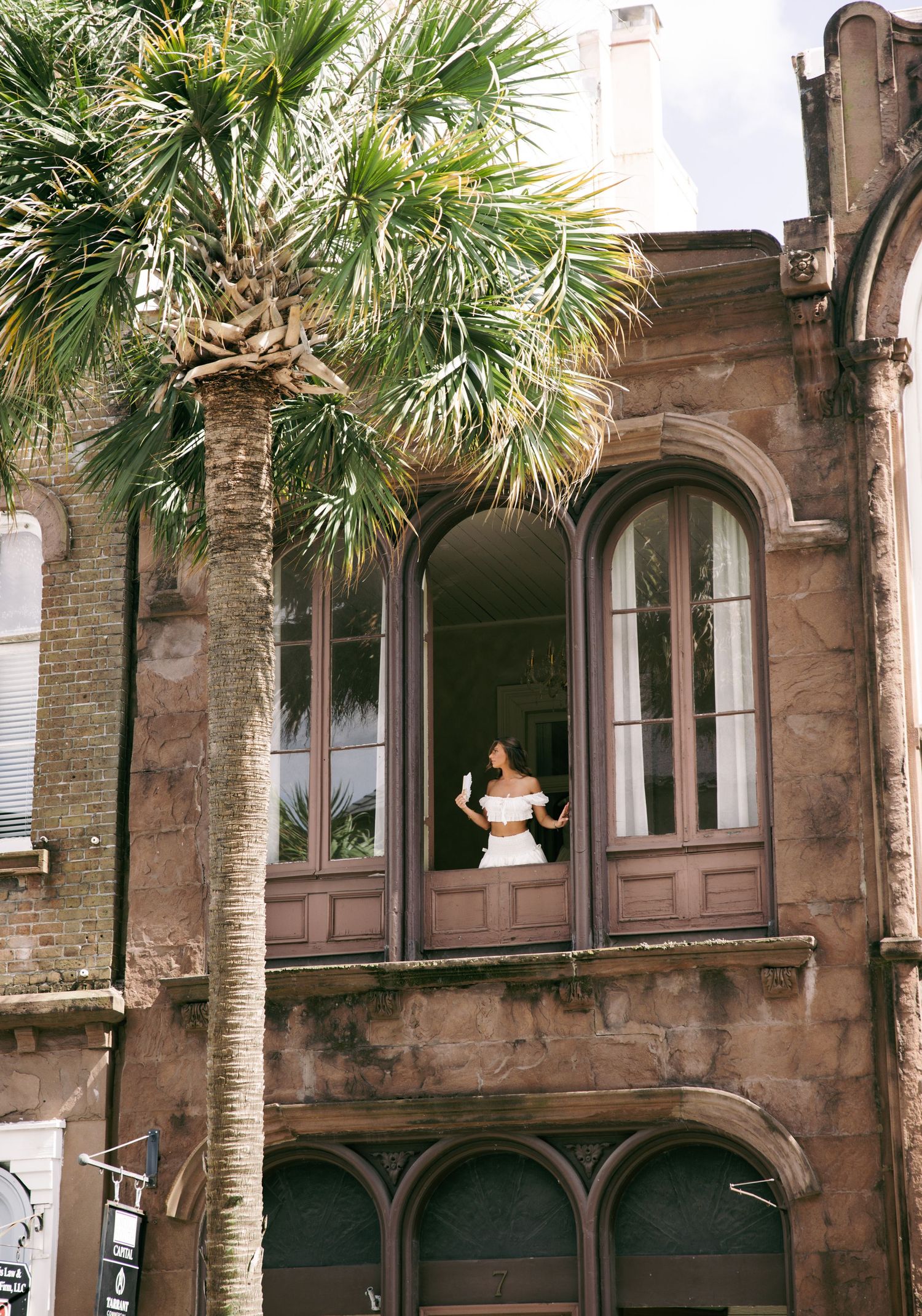 Woman in white top standing in arched window of historic stone building with palm tree in foreground.