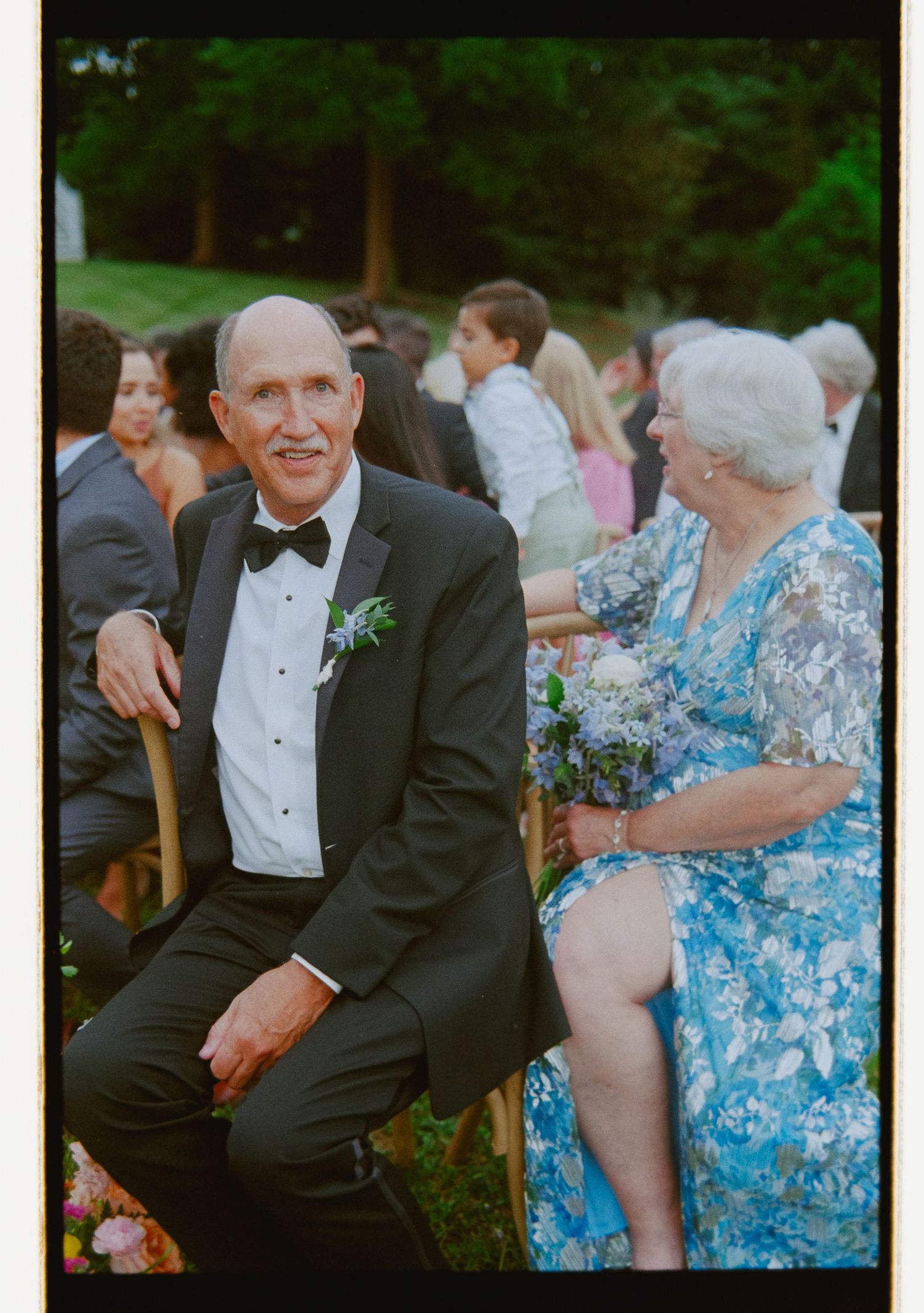 Smiling older gentleman in tuxedo sits with woman in blue floral dress at outdoor wedding ceremony.