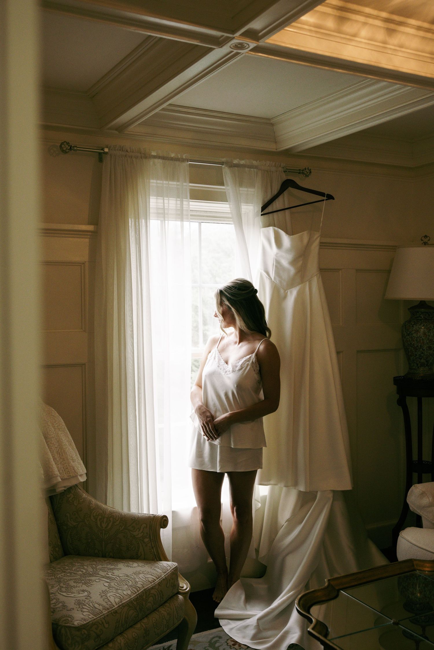 Bride in white slip gazes out window beside her elegant strapless wedding gown hanging in sunlit room.