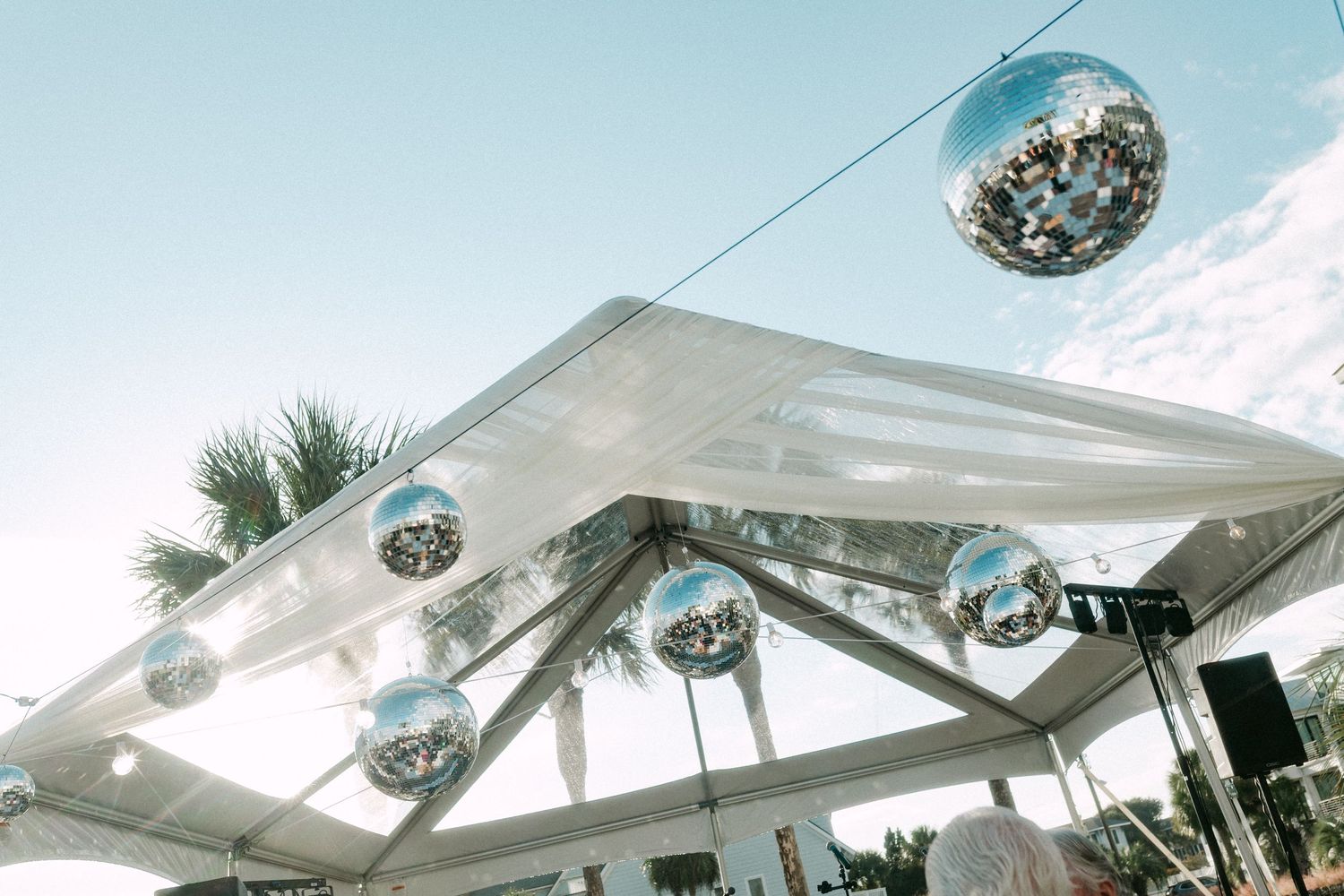 Silver disco balls hanging under a white tent canopy with palm trees and blue sky at an outdoor event.