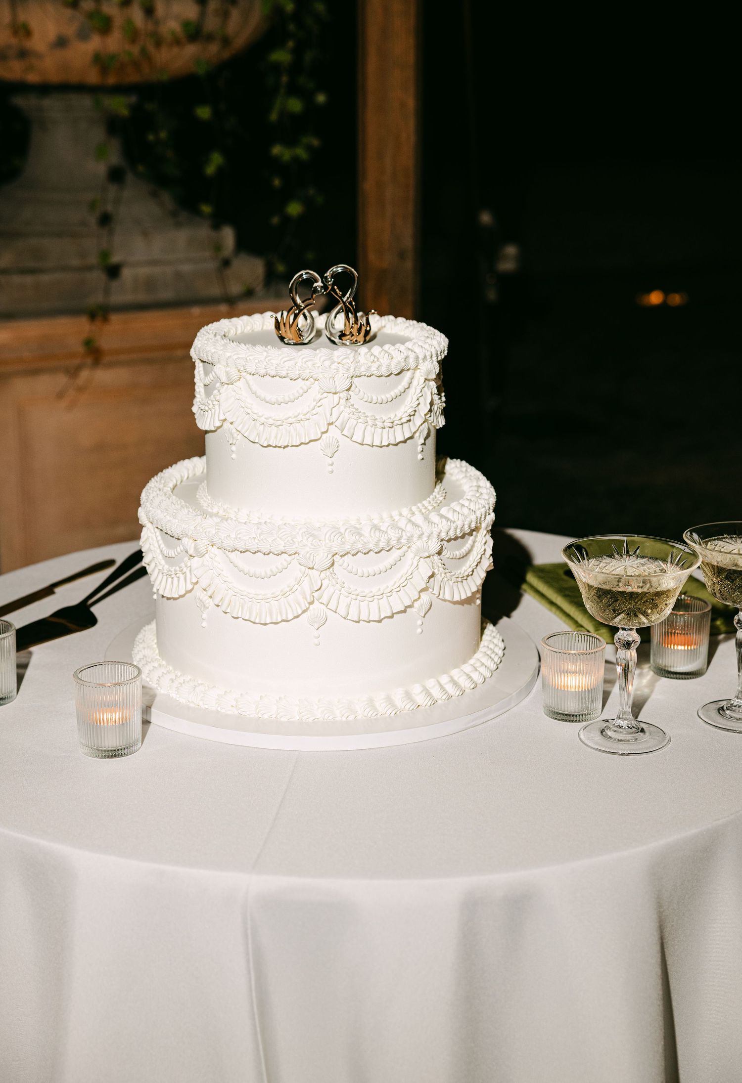 Two-tier white wedding cake with ruffle piping decoration and monogram topper on white linen table.
