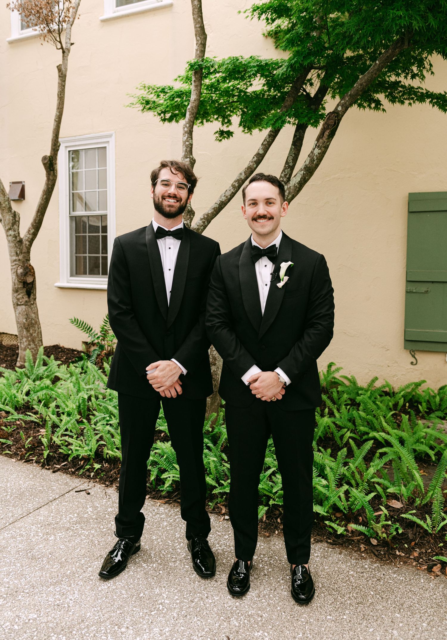 Two groomsmen in classic black tuxedos with bow ties posing outdoors in front of lush green trees.