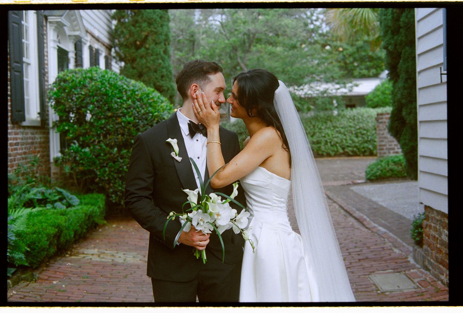 Bride and groom share tender kiss on brick path with white bouquet and veil.