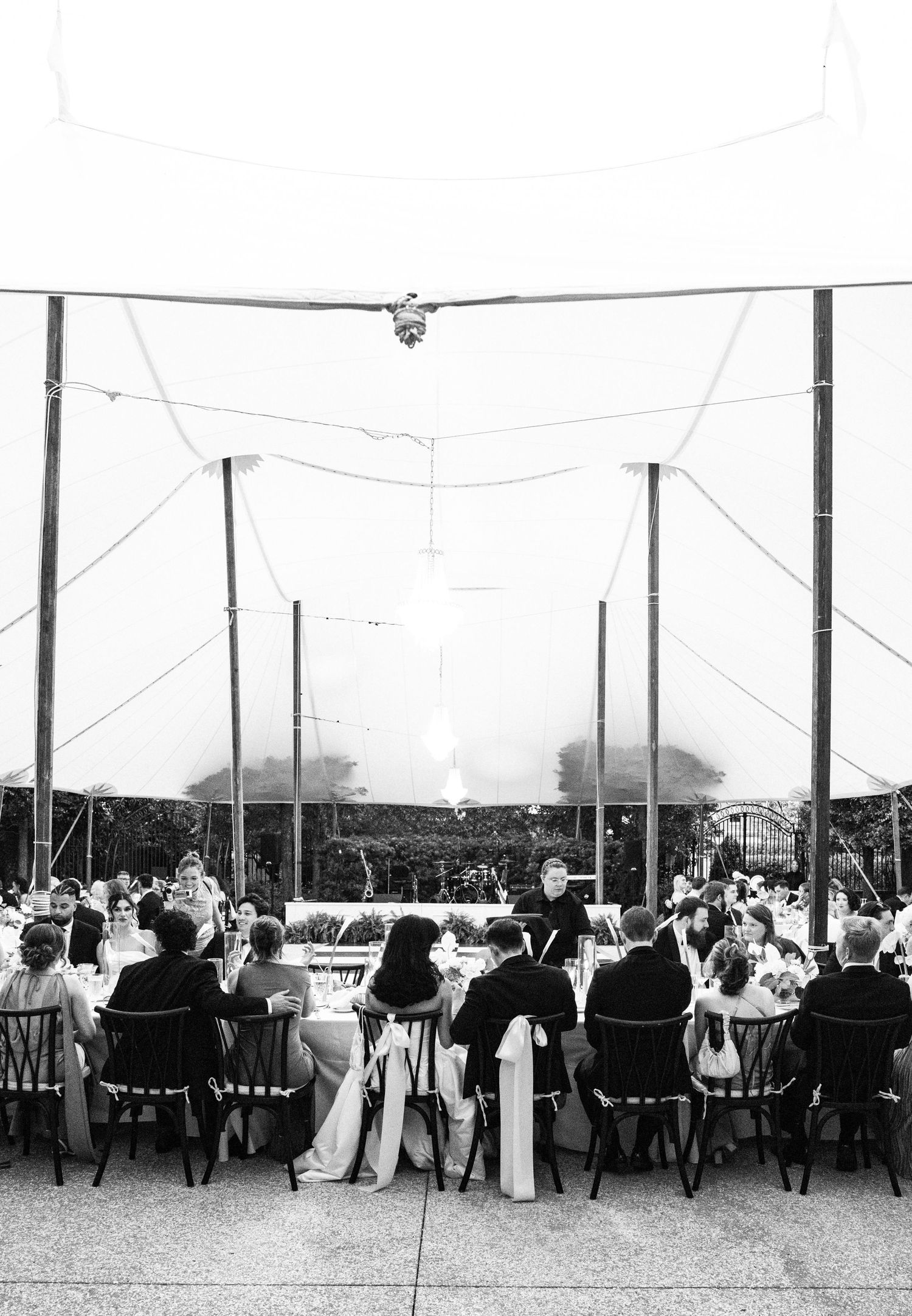 Black and white photo of wedding guests dining under a sailcloth tent with string lights overhead.