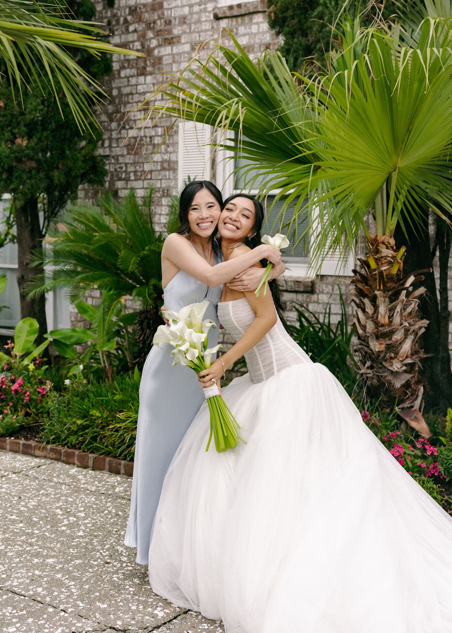 A bride in a white ballgown hugs her bridesmaid in blue while holding a white calla lily bouquet outdoors.