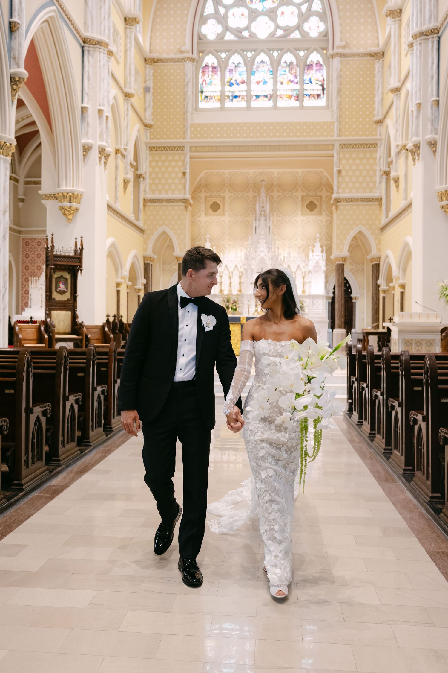 Newlyweds walk down ornate church aisle with stained glass windows and cream colored pillars.