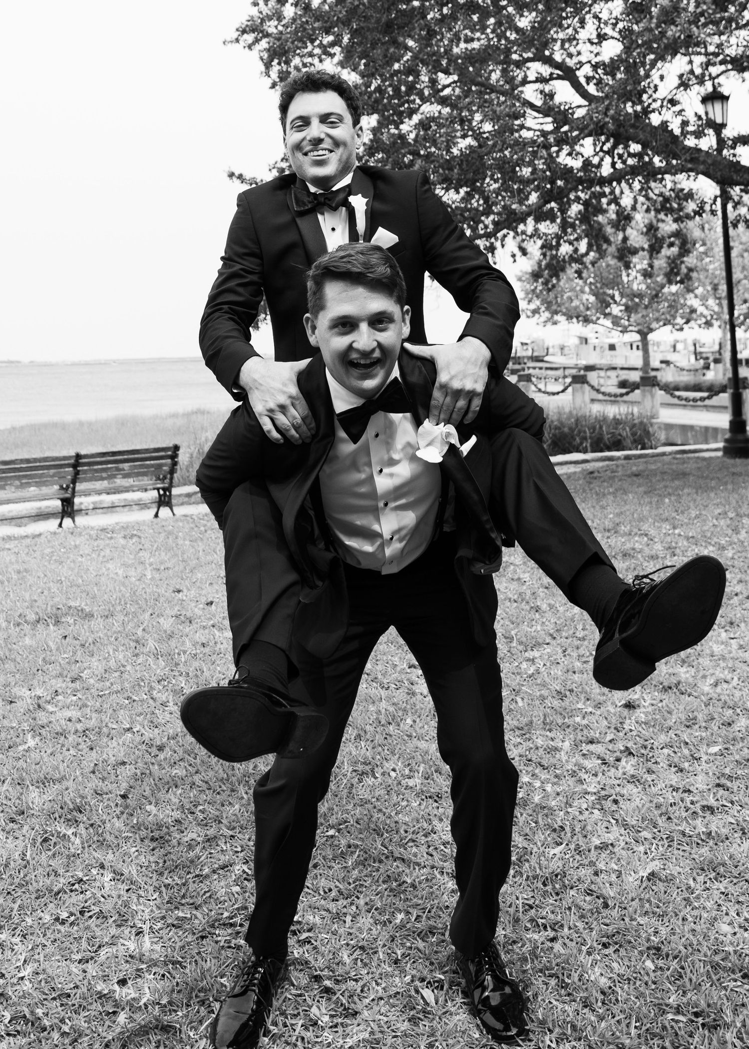 Groomsmen in black suits jump and pose playfully on grassy lawn during wedding photoshoot.