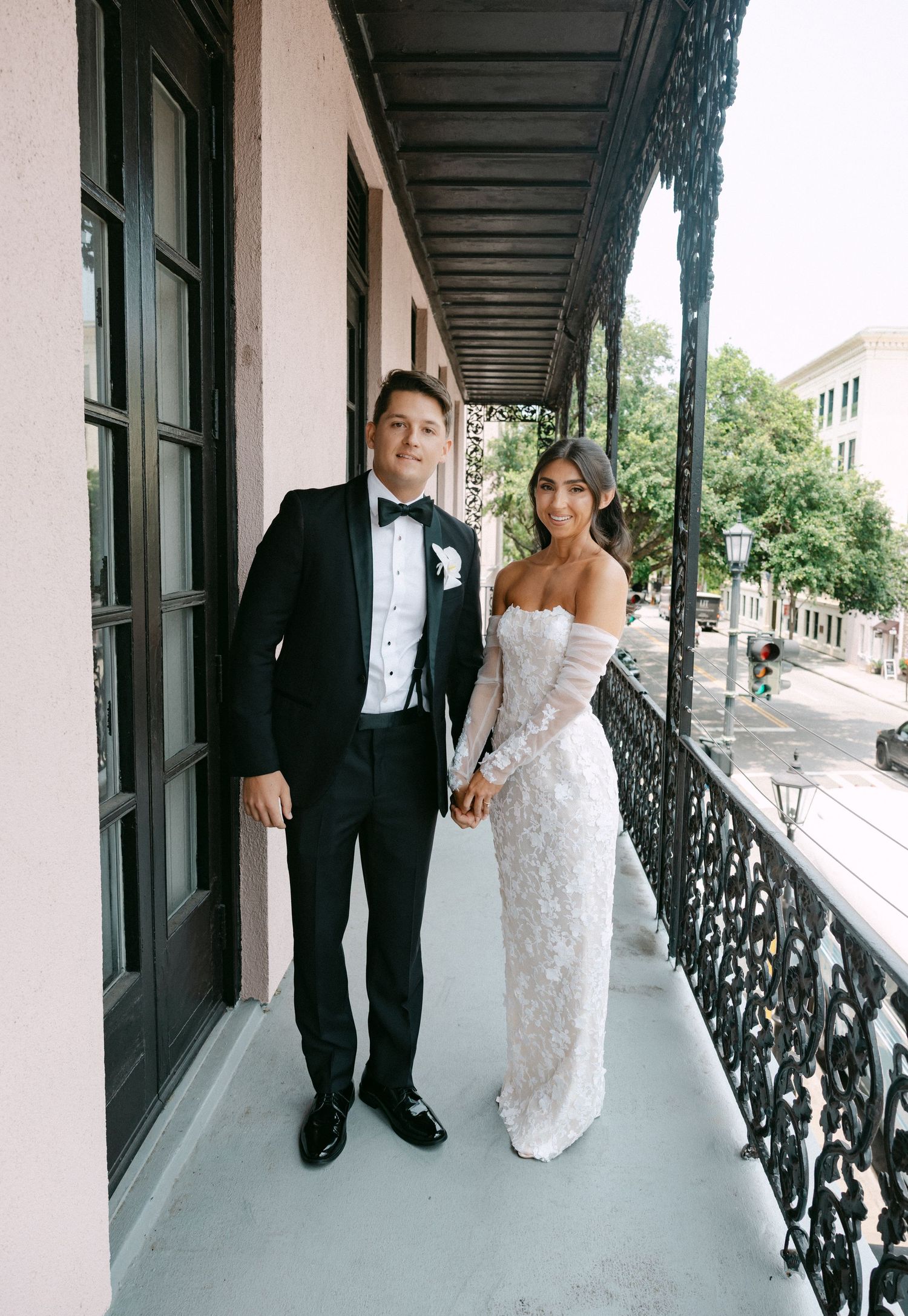 A wedding couple walks along an elegant covered porch with decorative black iron railings.