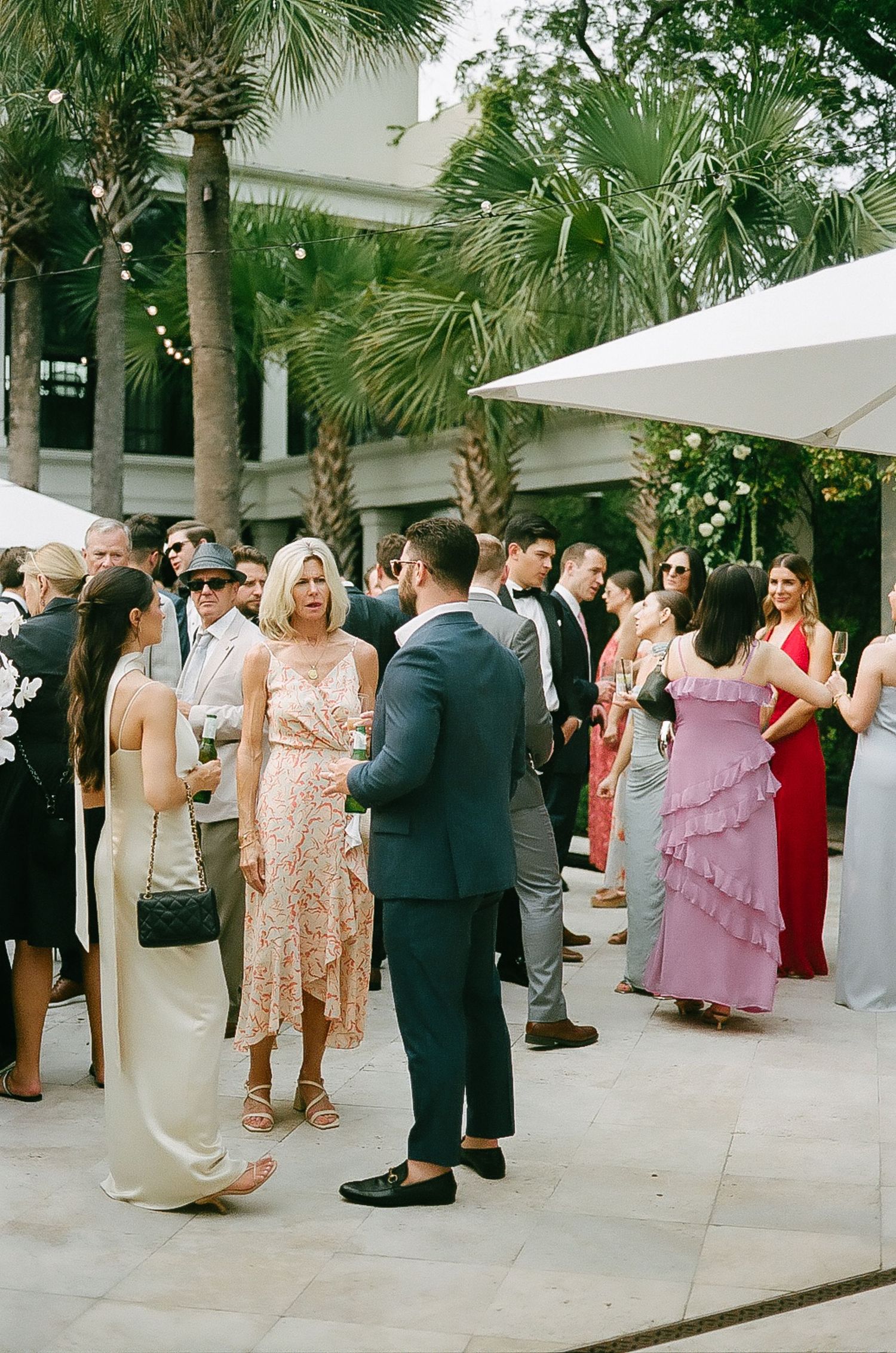 Wedding guests socializing on a patio surrounded by palm trees and white umbrellas.