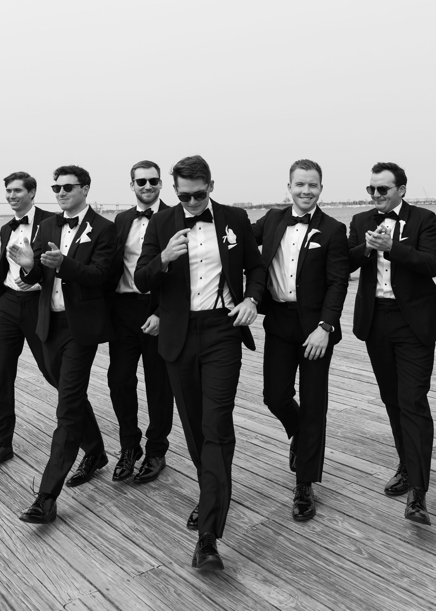Groomsmen in black tuxedos strike playful poses on a wooden boardwalk.