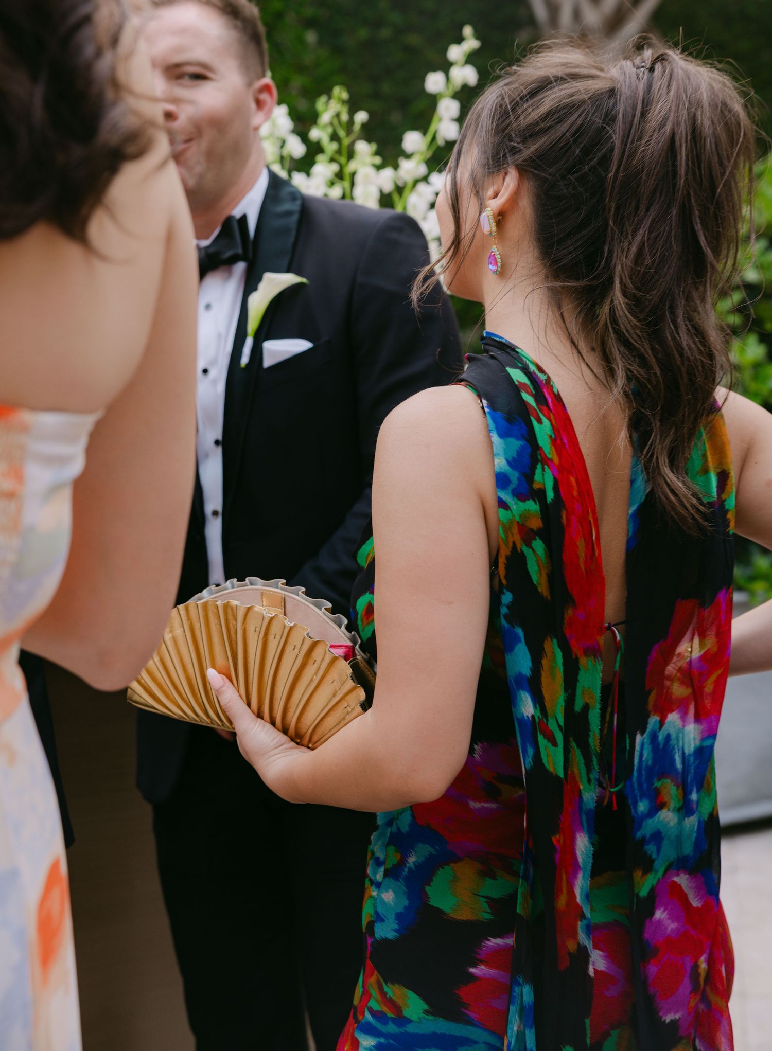 Wedding guests chatting outdoors, with a colorful floral dress and formal tuxedo visible.