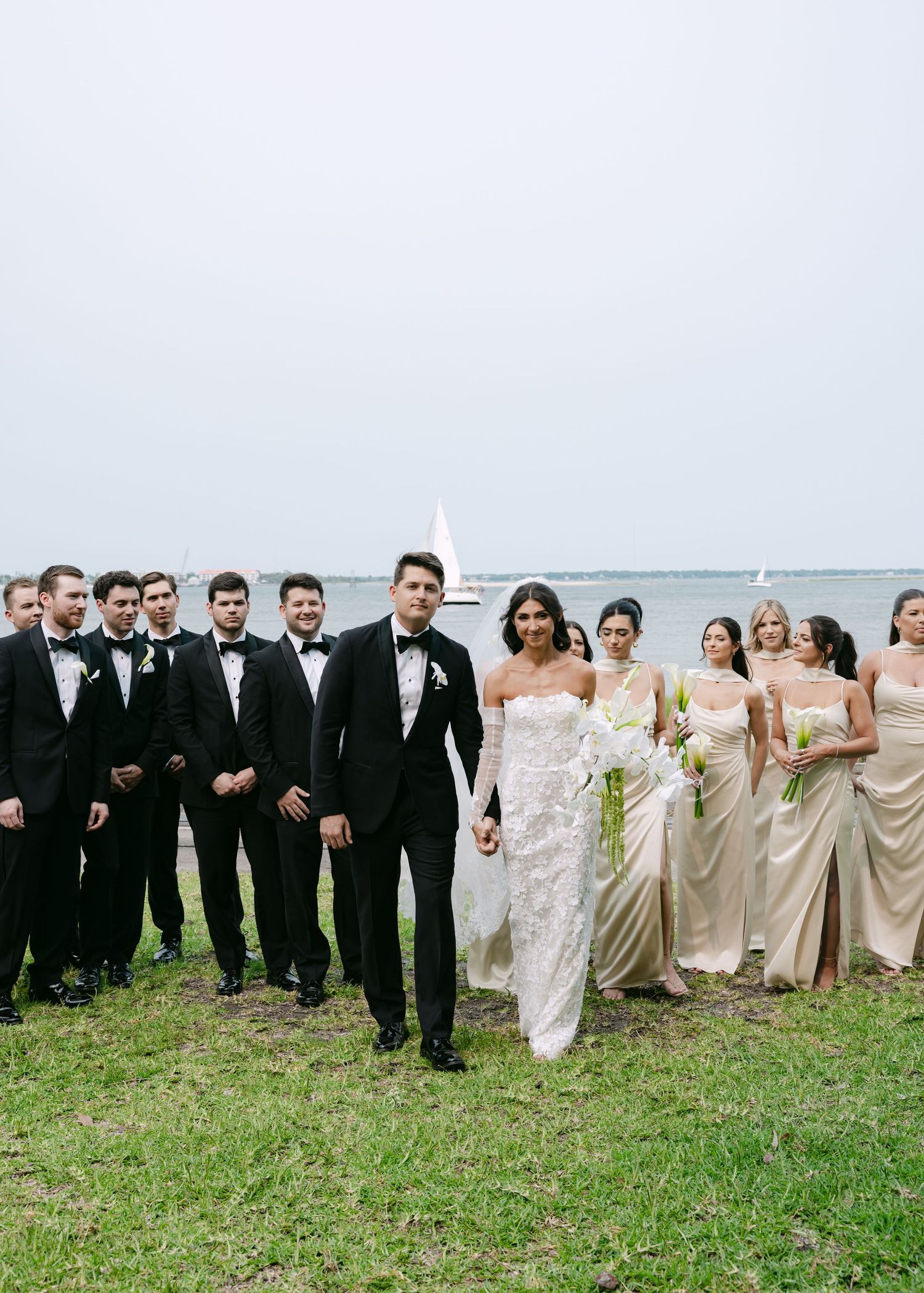 Wedding party with groomsmen in black tuxedos and bridesmaids in champagne dresses pose outdoors.