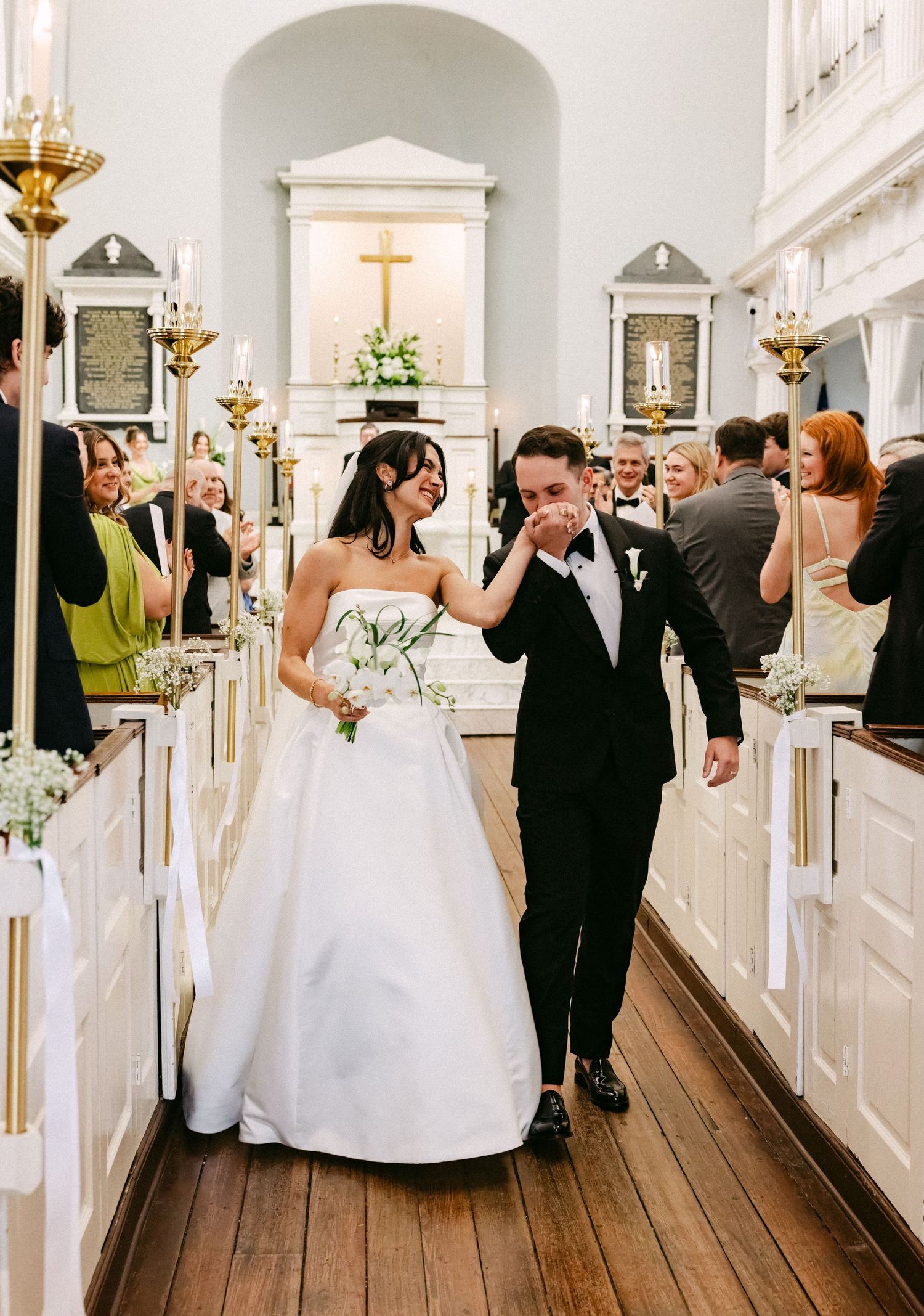 Bride and groom walk down church aisle after wedding ceremony, bride wiping groom's tear, guests watching.