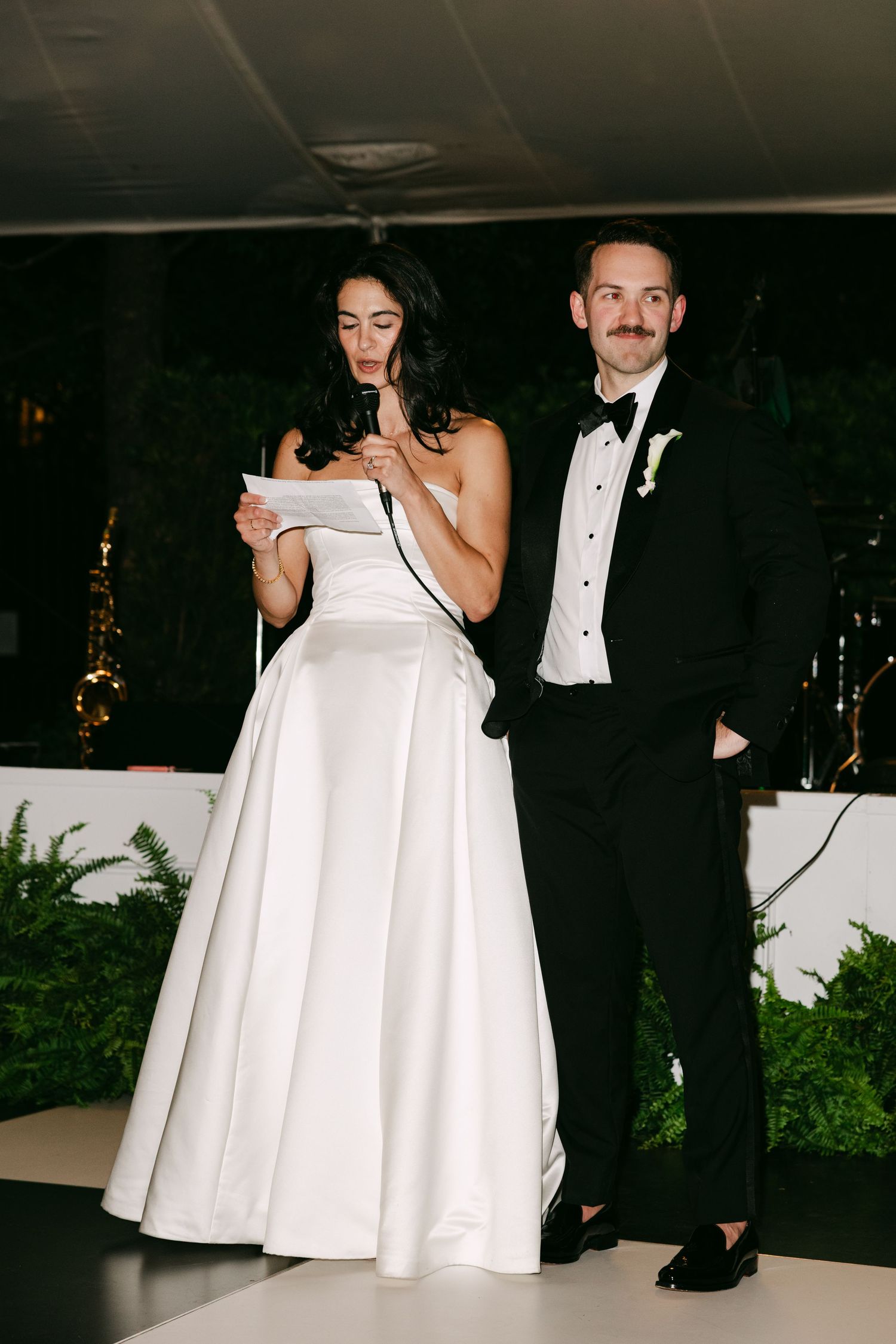 Bride in strapless white ball gown reads speech at microphone beside groom in black tuxedo at wedding reception.
