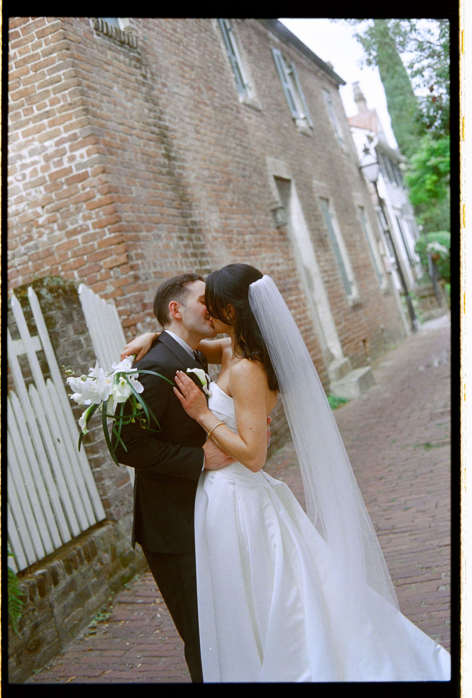 Bride and groom sharing a kiss on historic brick street with wedding veil flowing.