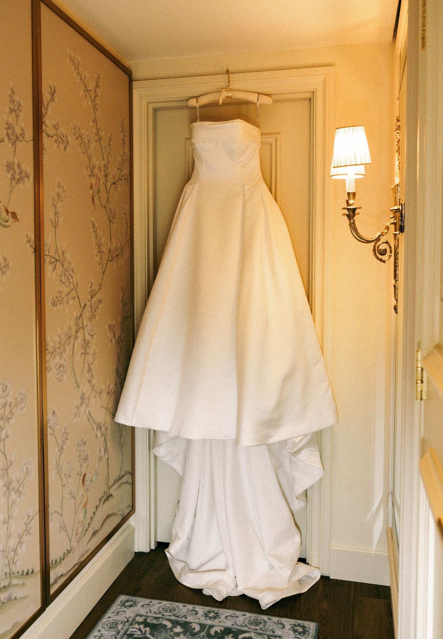 Strapless white ball gown wedding dress hanging in an elegantly decorated room with floral wallpaper.