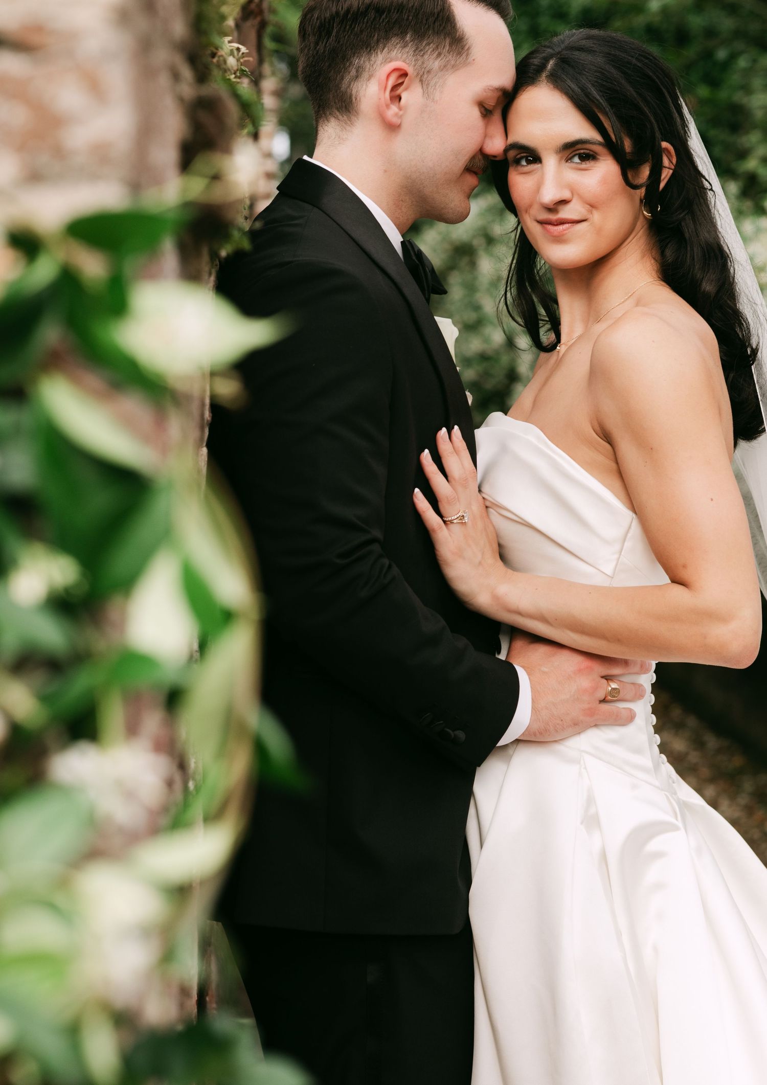 Bride in white strapless gown embraces groom in black tuxedo outdoors, surrounded by lush greenery.