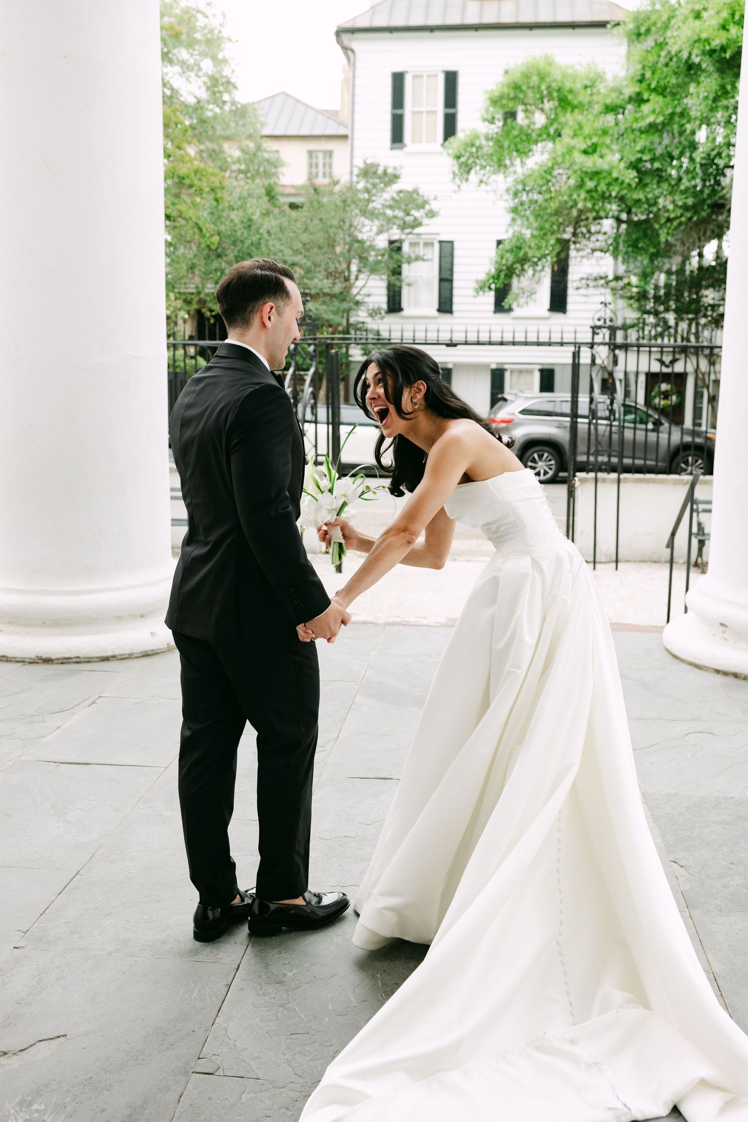Bride in strapless white gown holds grooms hand outside historic venue with columns.