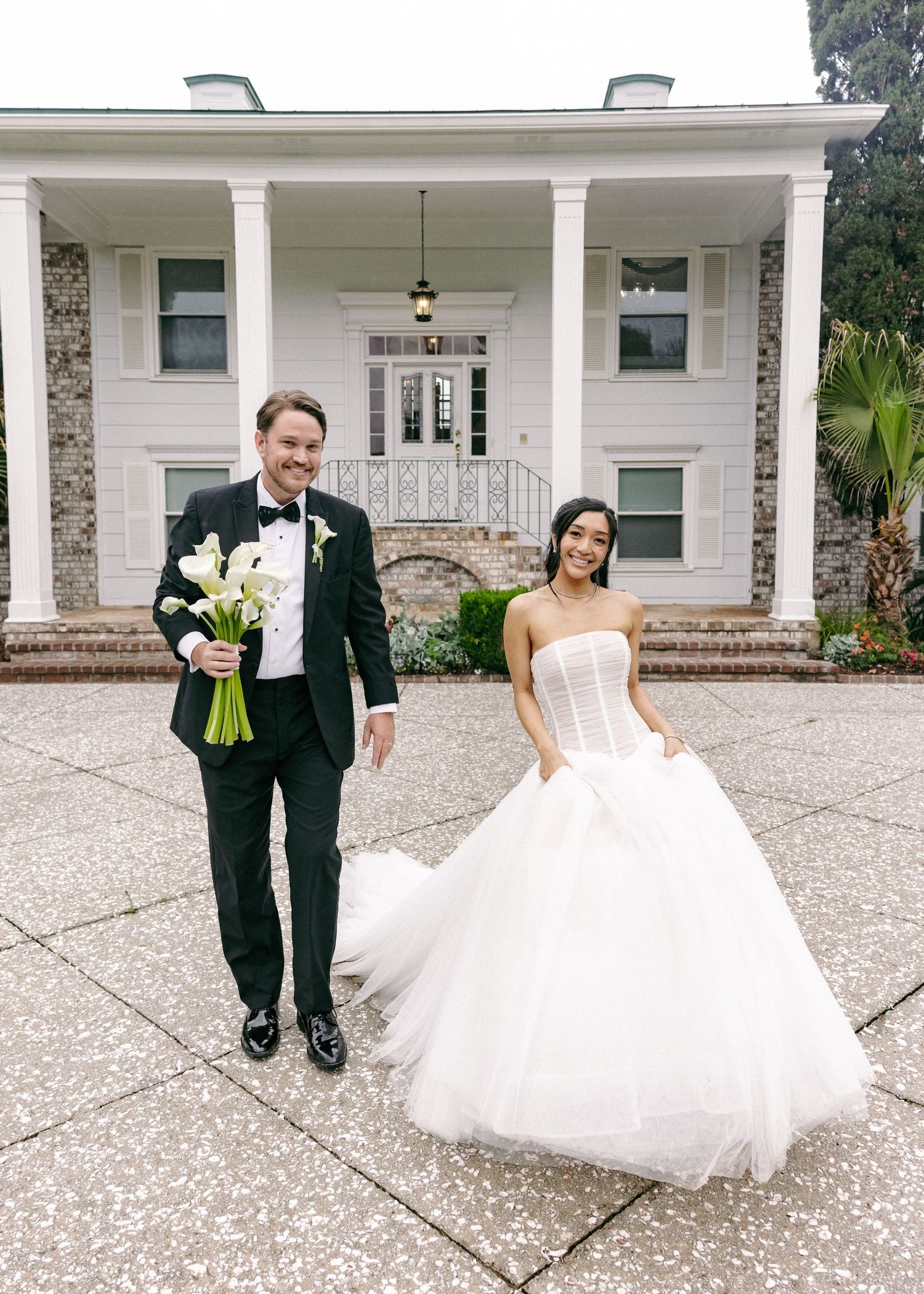 Smiling bride and groom pose outside a classic white columned estate after their wedding ceremony.