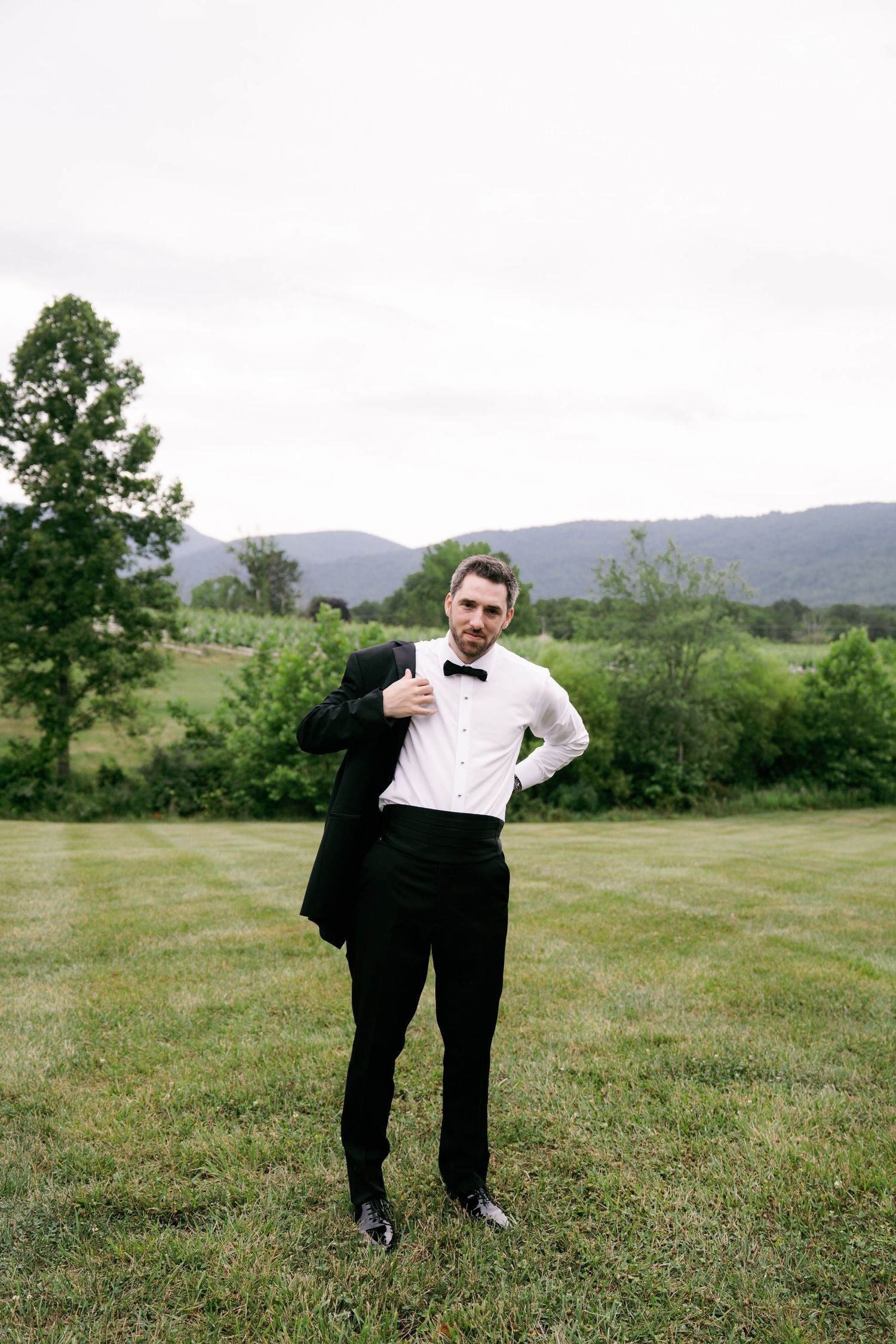 Groom in black tuxedo and bow tie stands on green lawn with mountain backdrop.