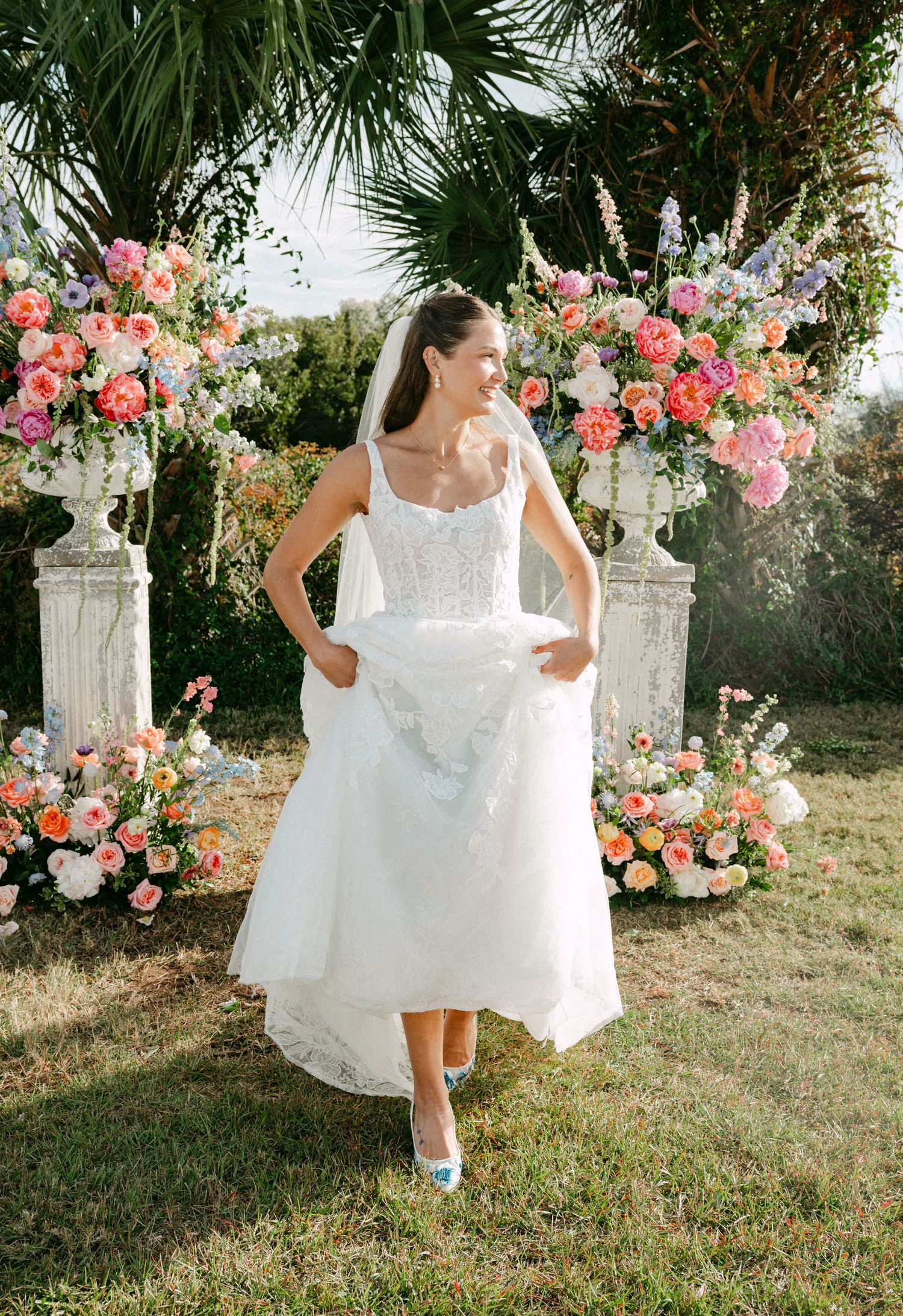 Smiling bride in lace gown and veil lifting dress hem before colorful floral arch outdoors.