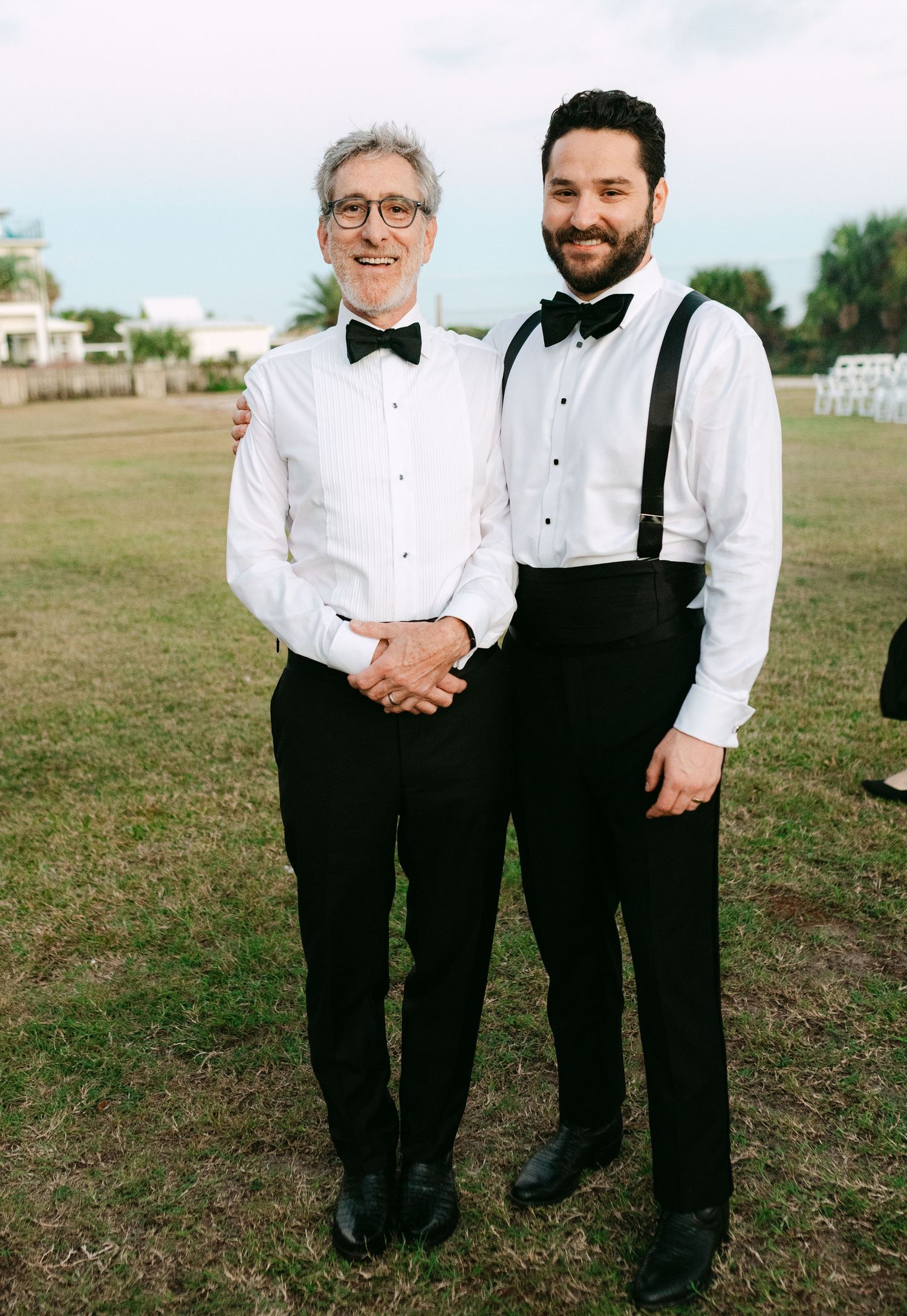Two men in formal black tuxedos with bow ties posing outdoors at wedding.