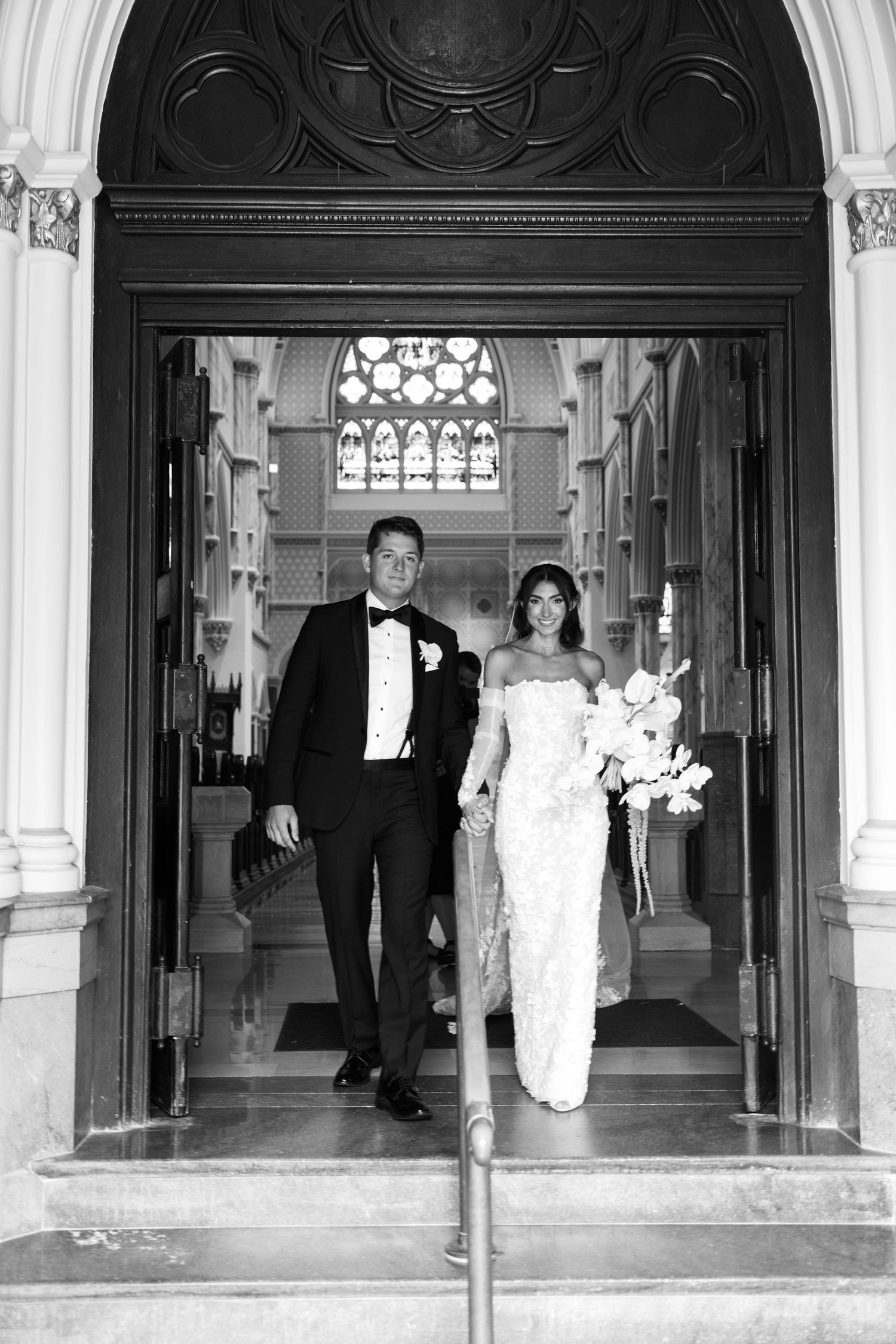 Elegant black and white wedding photos taken in a grand hallway with ornate gothic architecture and stained glass windows.