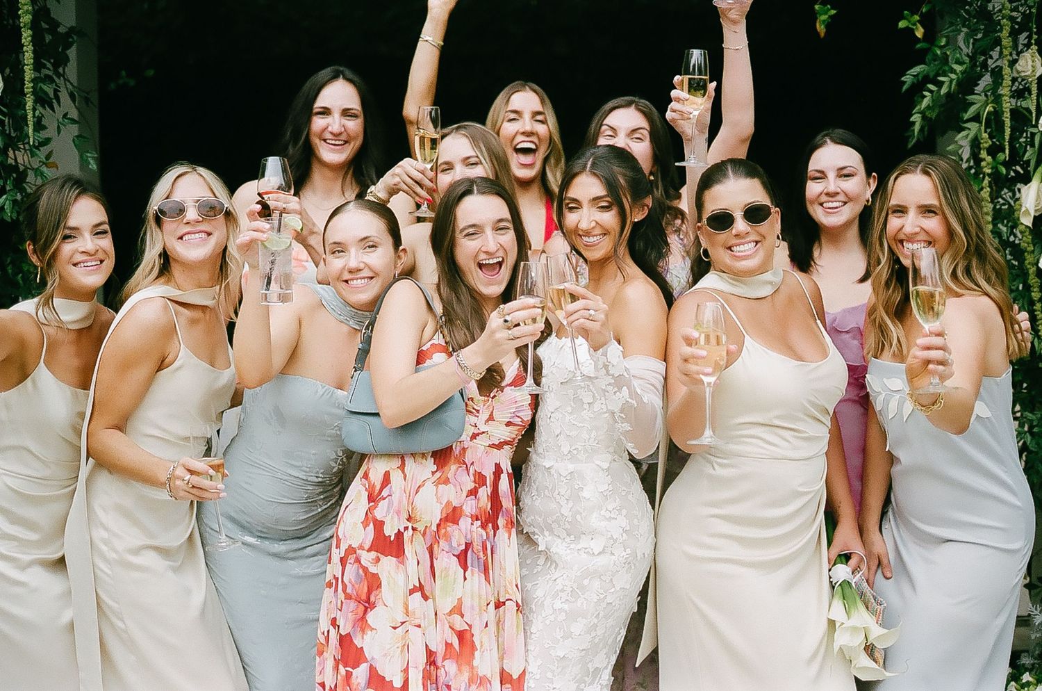 Wedding party of bridesmaids in coordinated dresses pose together at outdoor celebration.