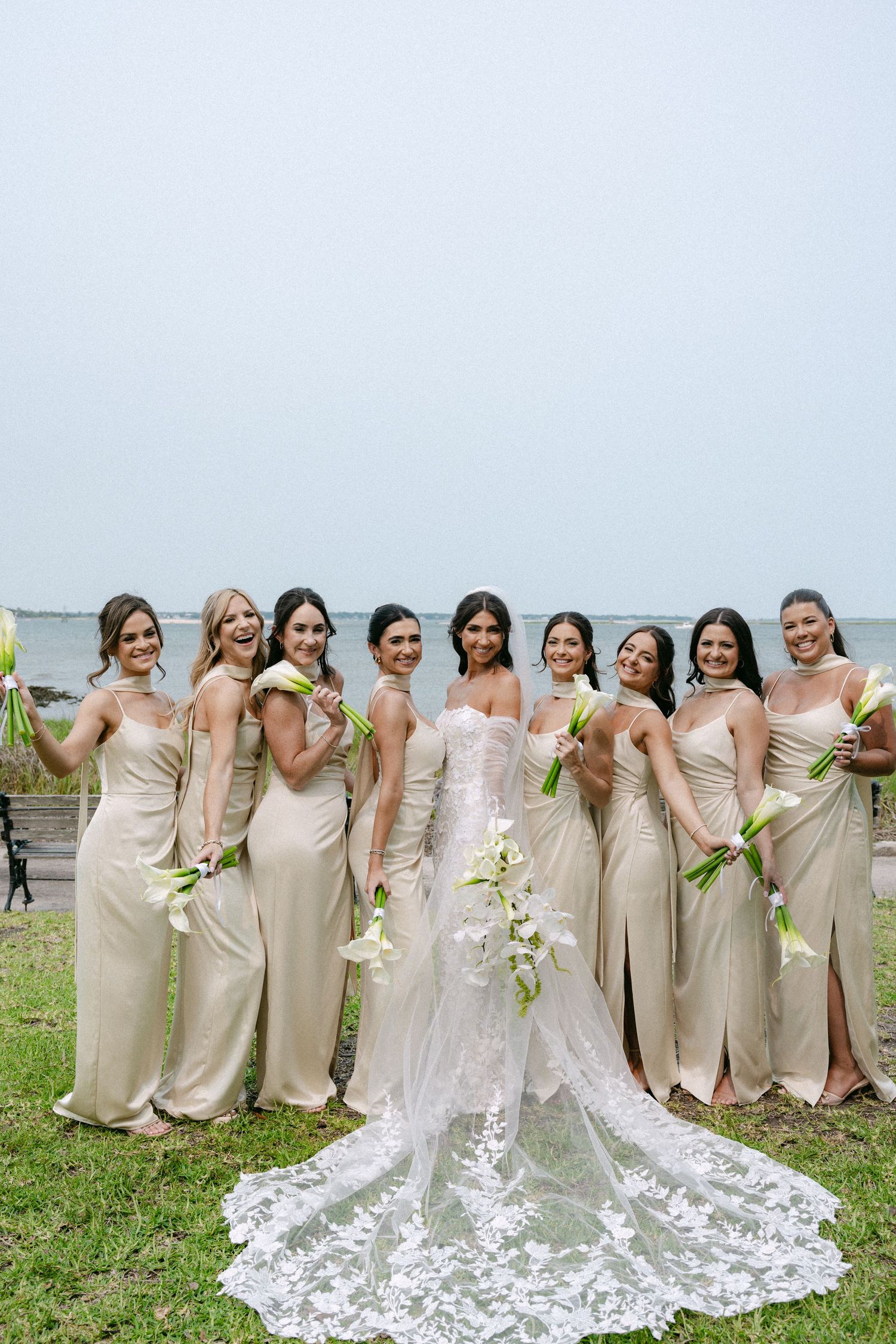 A bridal party in matching champagne dresses poses in a line on grass near the ocean.