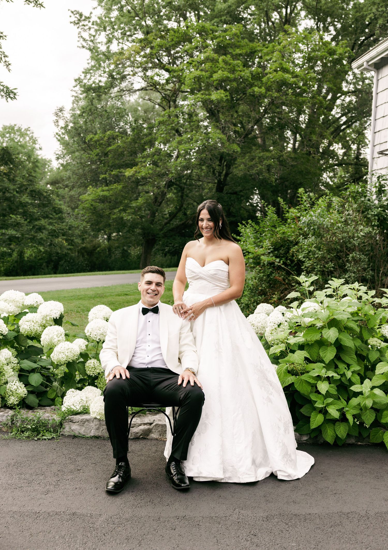 Bride and groom pose among white hydrangeas, groom seated in chair wearing ivory tuxedo jacket.