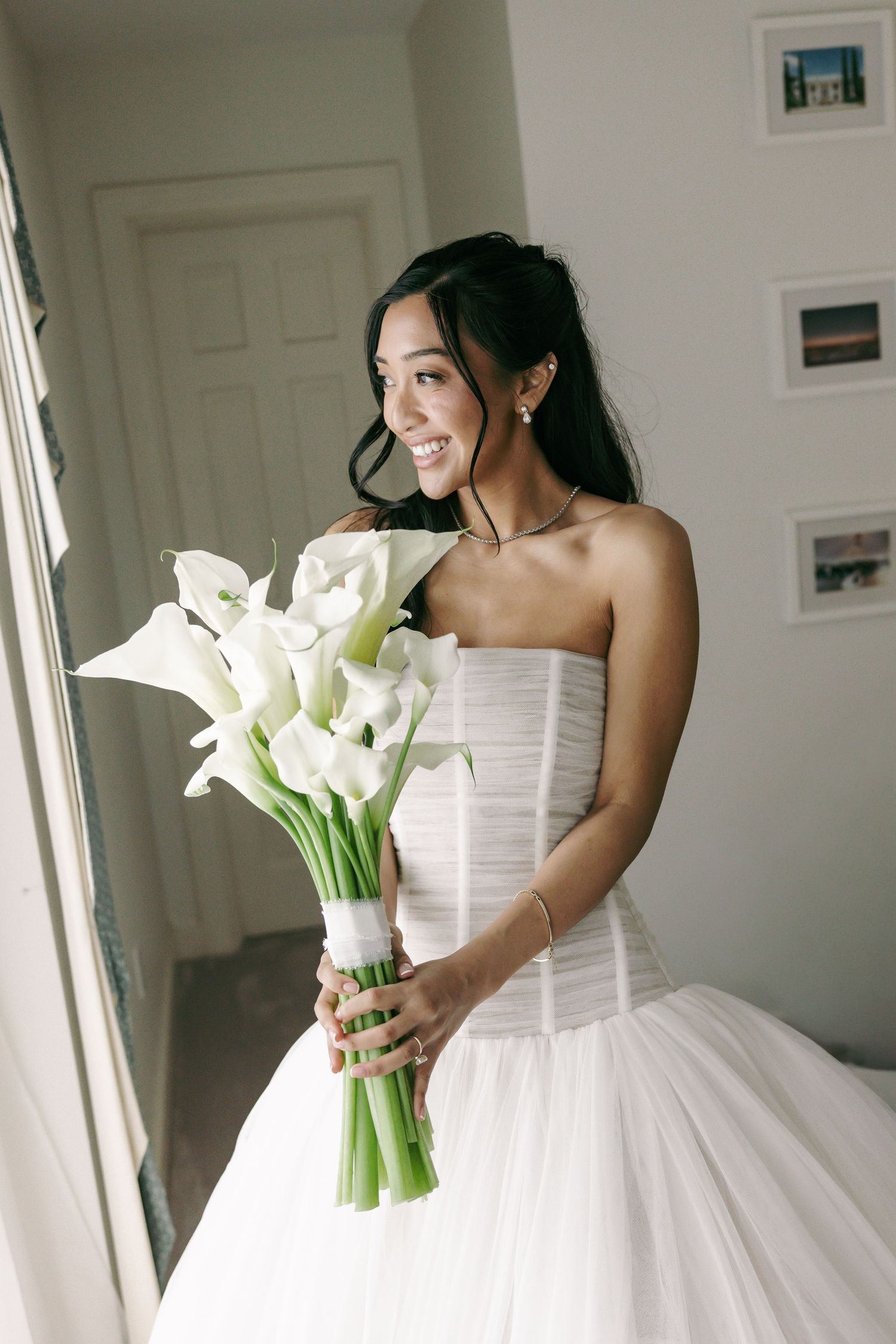 Smiling bride in strapless white gown holding a bouquet of white calla lilies near a window.