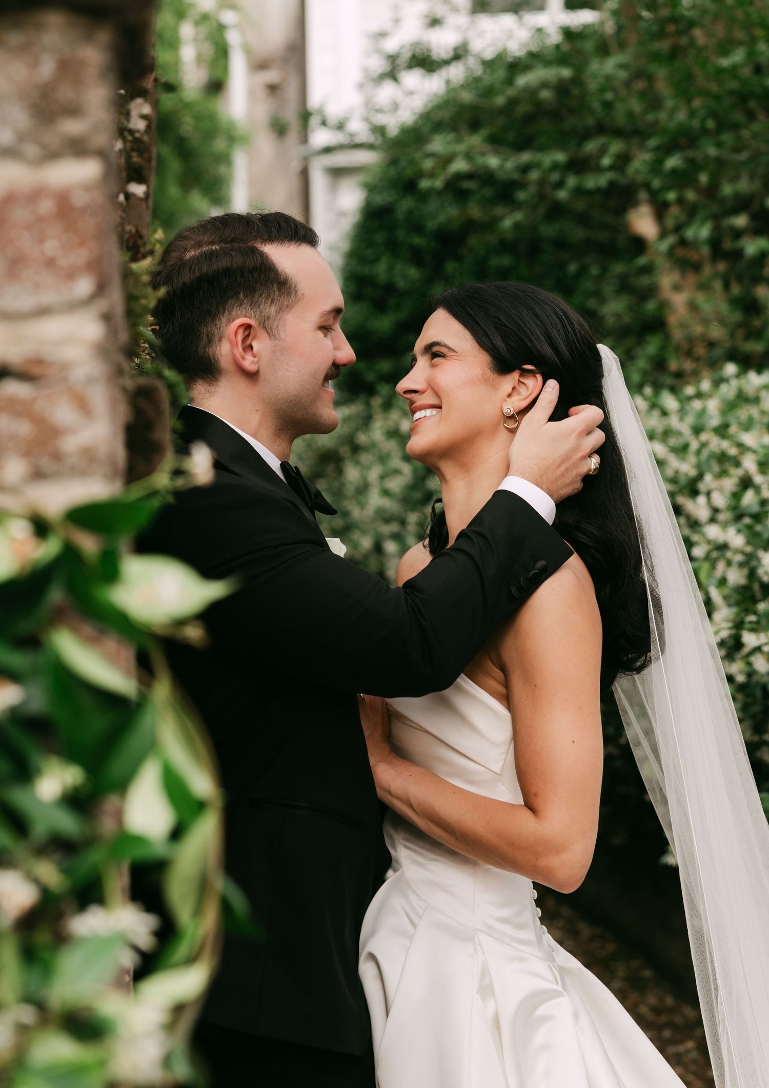 Smiling bride in white strapless gown and veil gazes at groom in black suit surrounded by lush greenery.