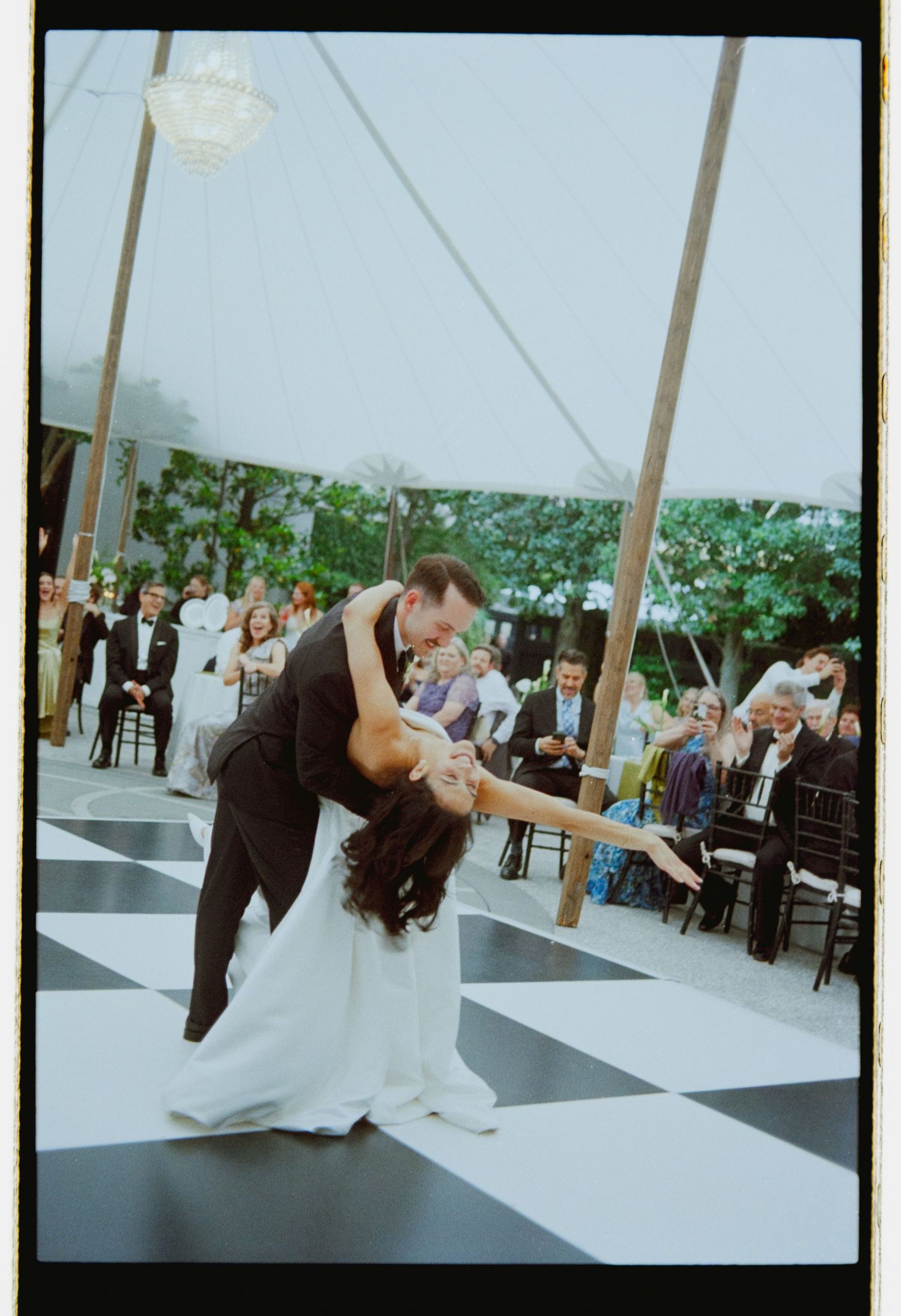 Groom dips bride on a black and white checkered dance floor during their first dance under a tent.