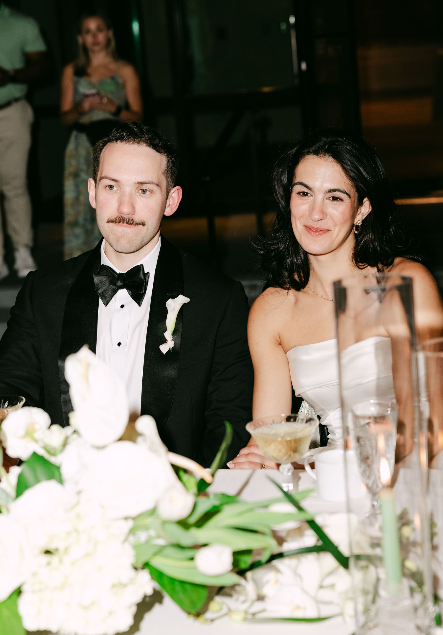 Bride and groom smile at wedding reception table with white floral centerpiece.