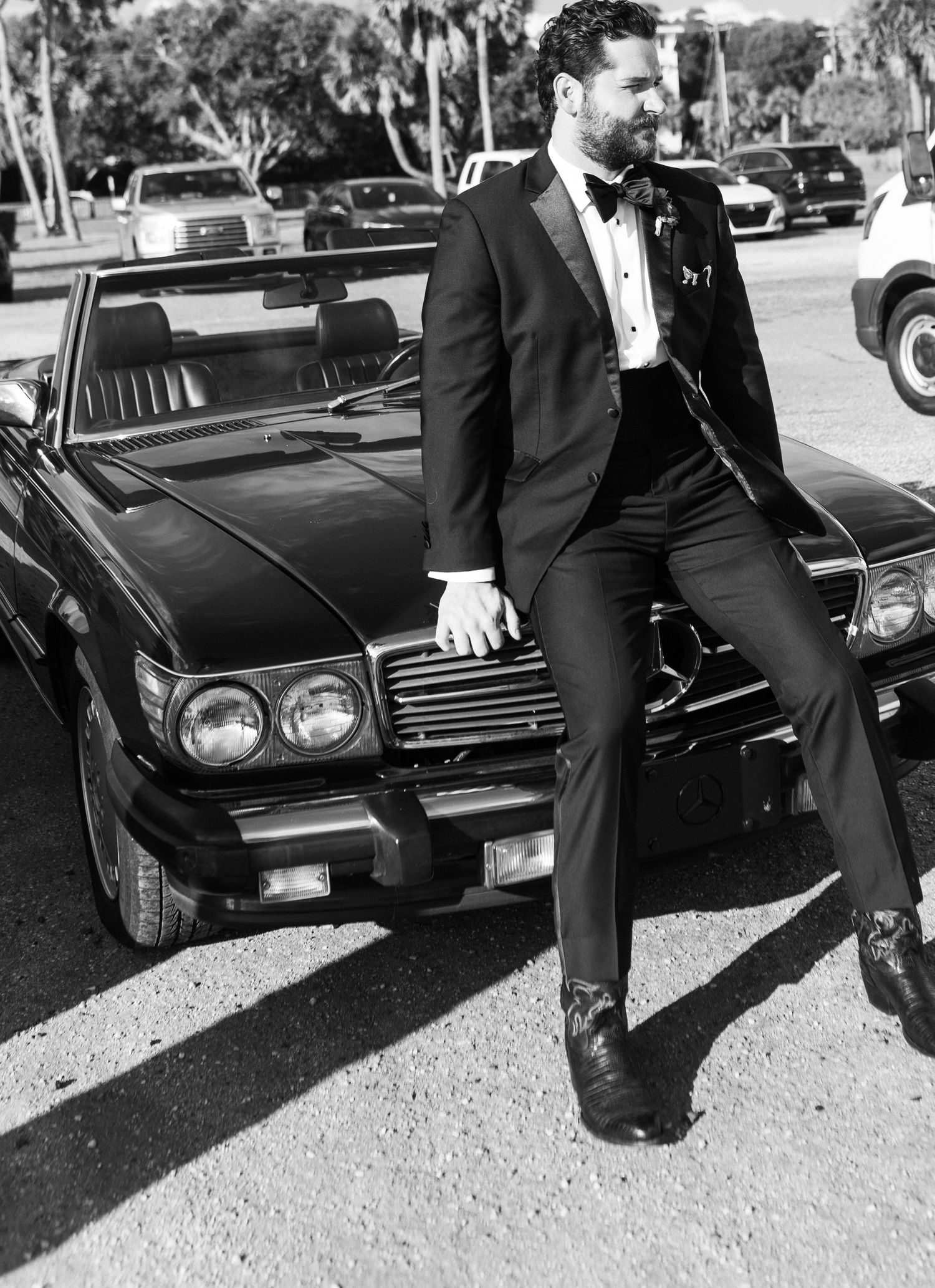Dapper groom in black tuxedo leaning against a vintage convertible car in black and white.