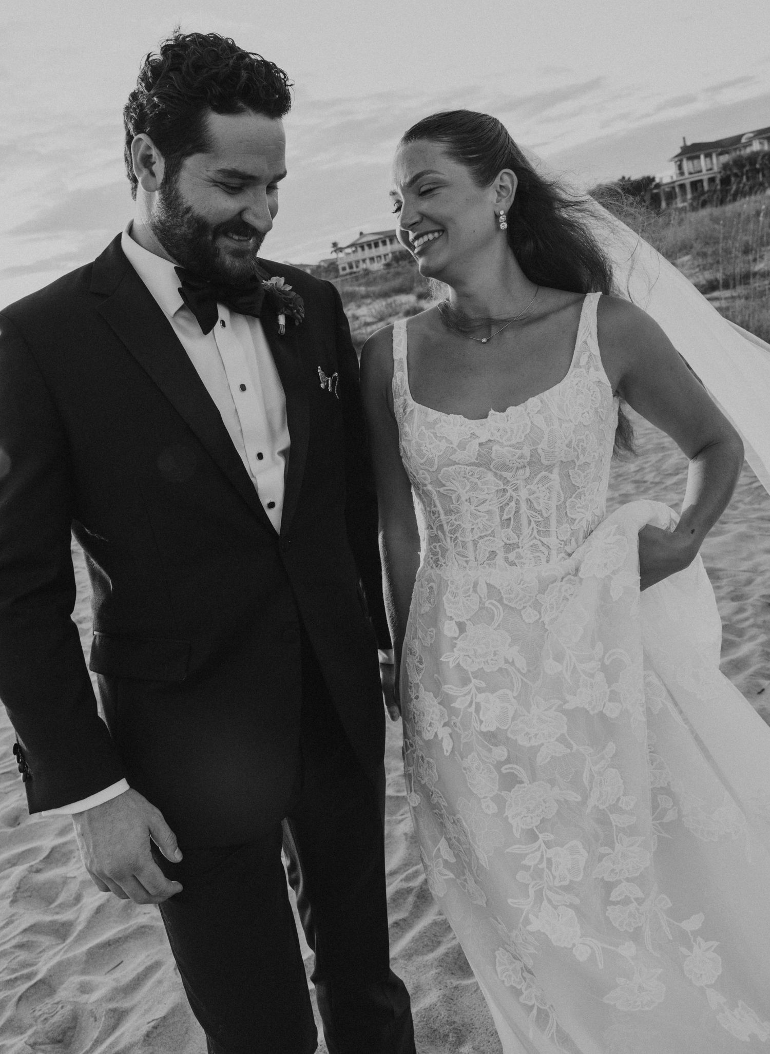 Black and white beach photo of smiling bride in lace gown and groom in tuxedo at dusk.