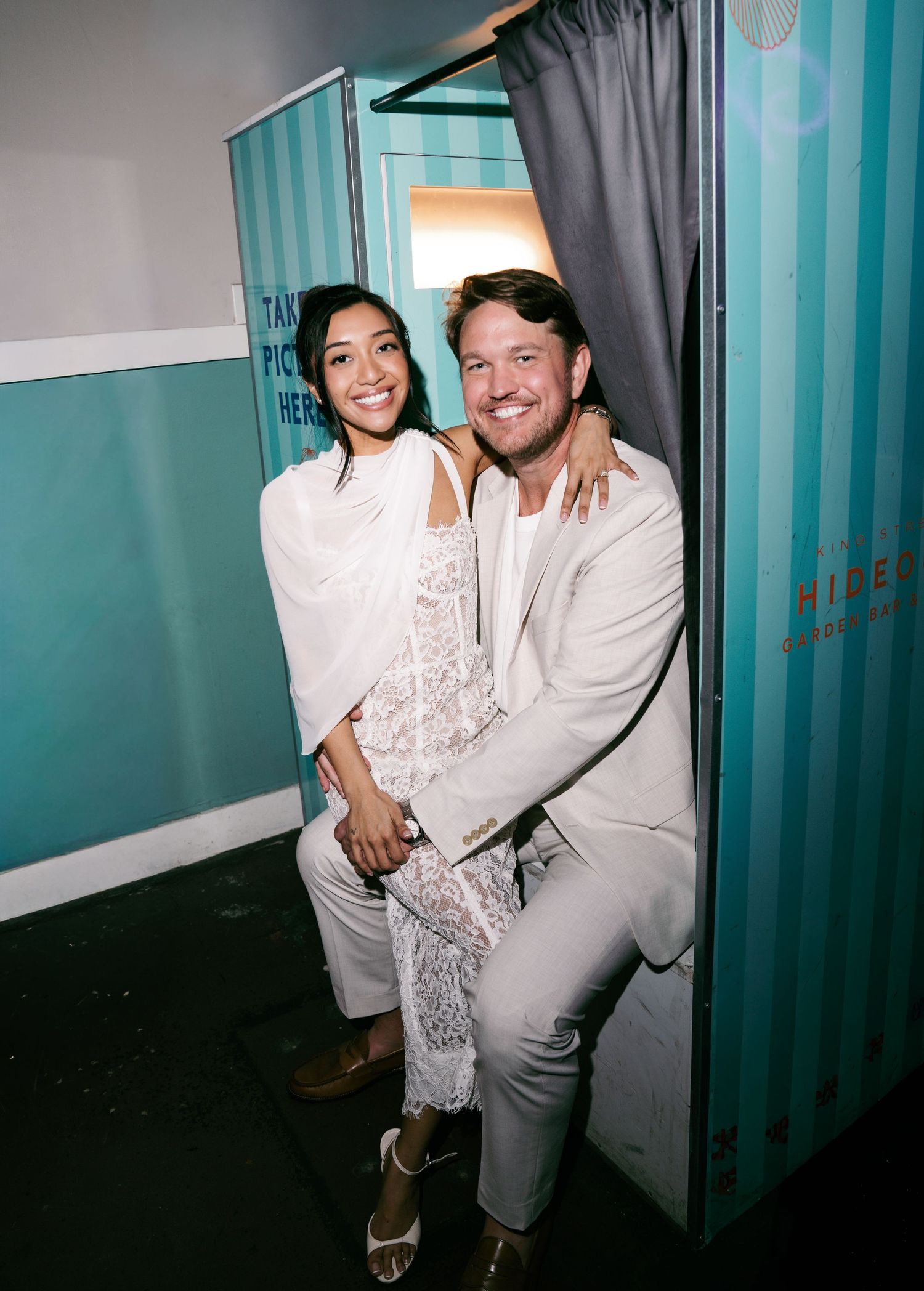Couple in white outfits smiling inside a teal photo booth, posing together for a fun photo.