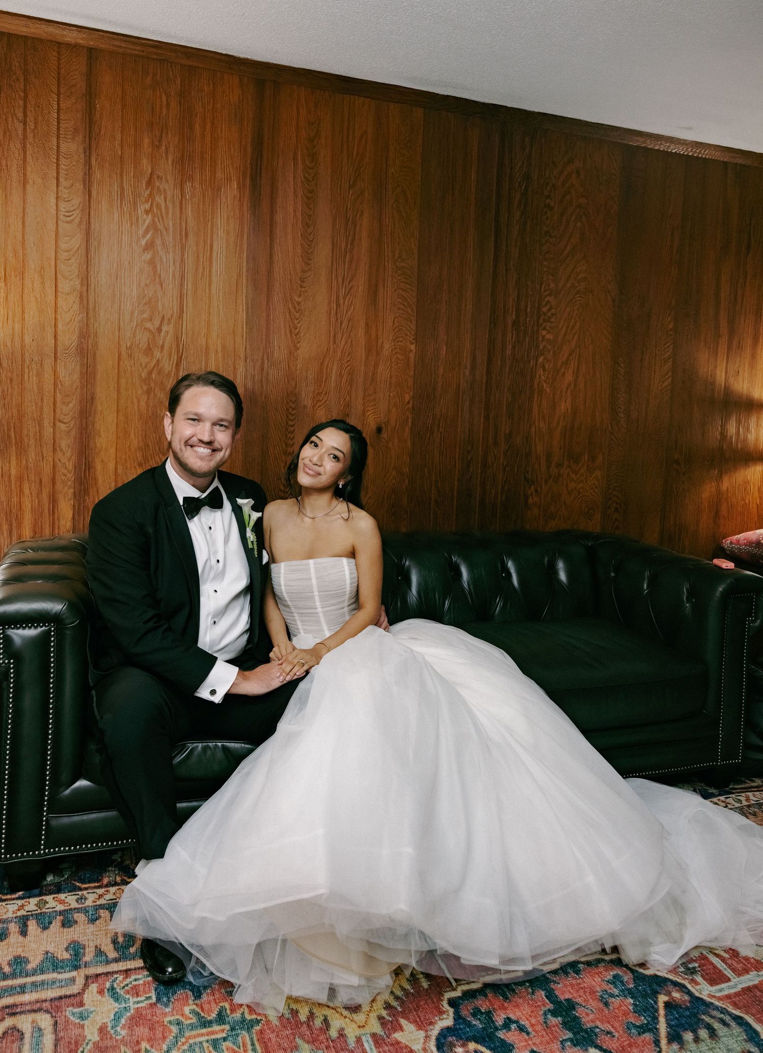 Bride in white ballgown and groom in black tuxedo sitting together on a green leather sofa at their wedding.