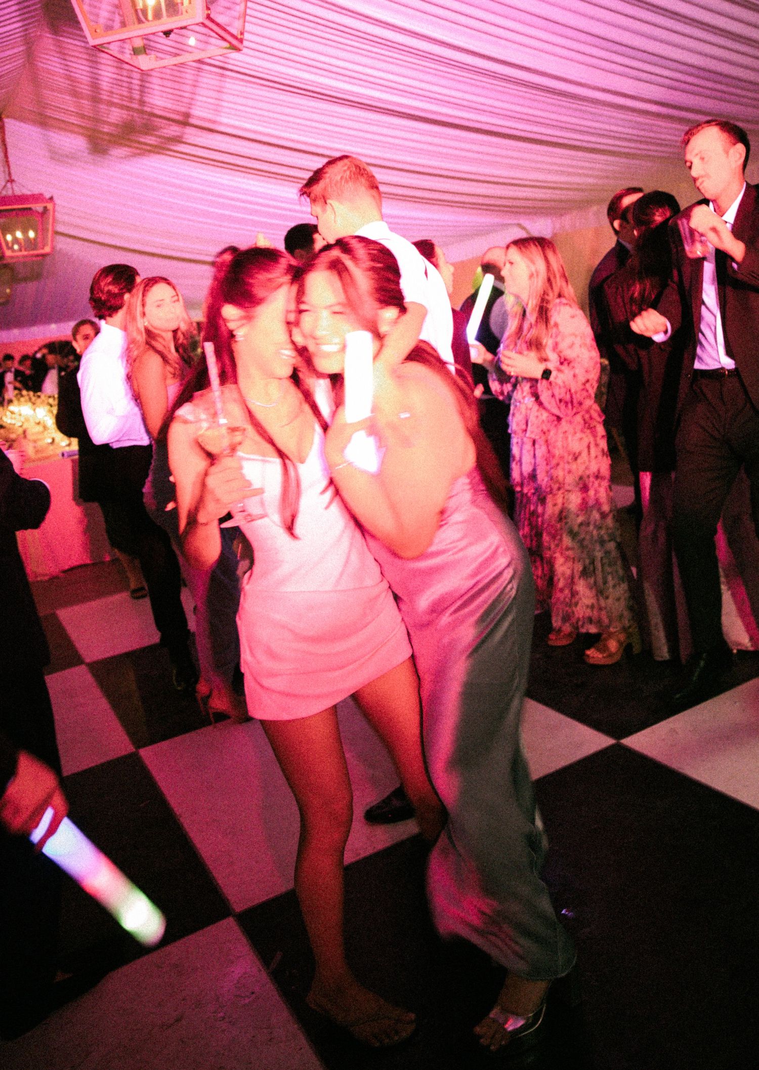 Guests dance joyfully on checkered floor at wedding reception under pink-lit tent.